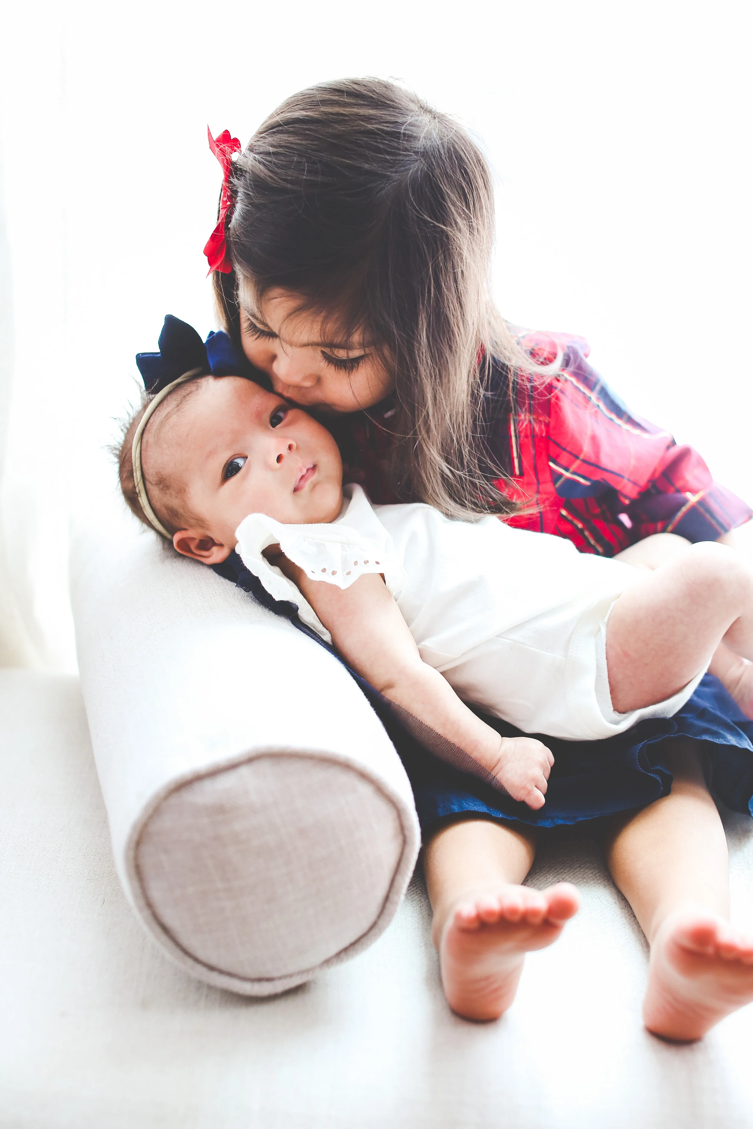 Portrait of a young big sister holding her infant baby sister on her lap & giving her a kiss on the cheek with bright lighting.