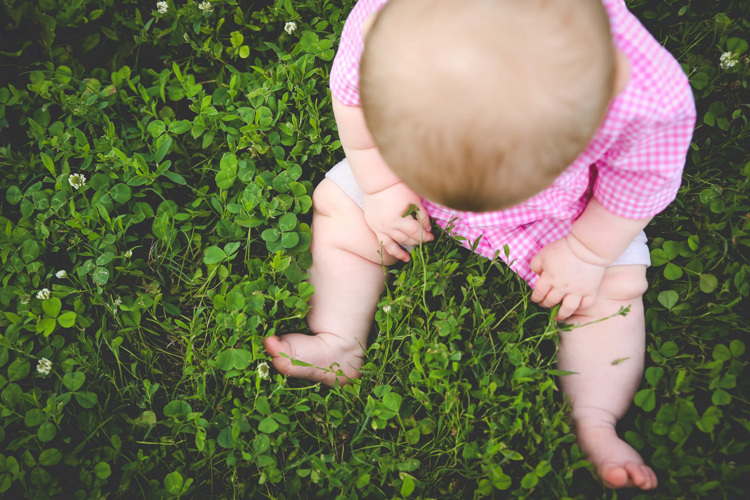 Bird's eye view of a baby's chunky legs and hands while sitting on green grass.
