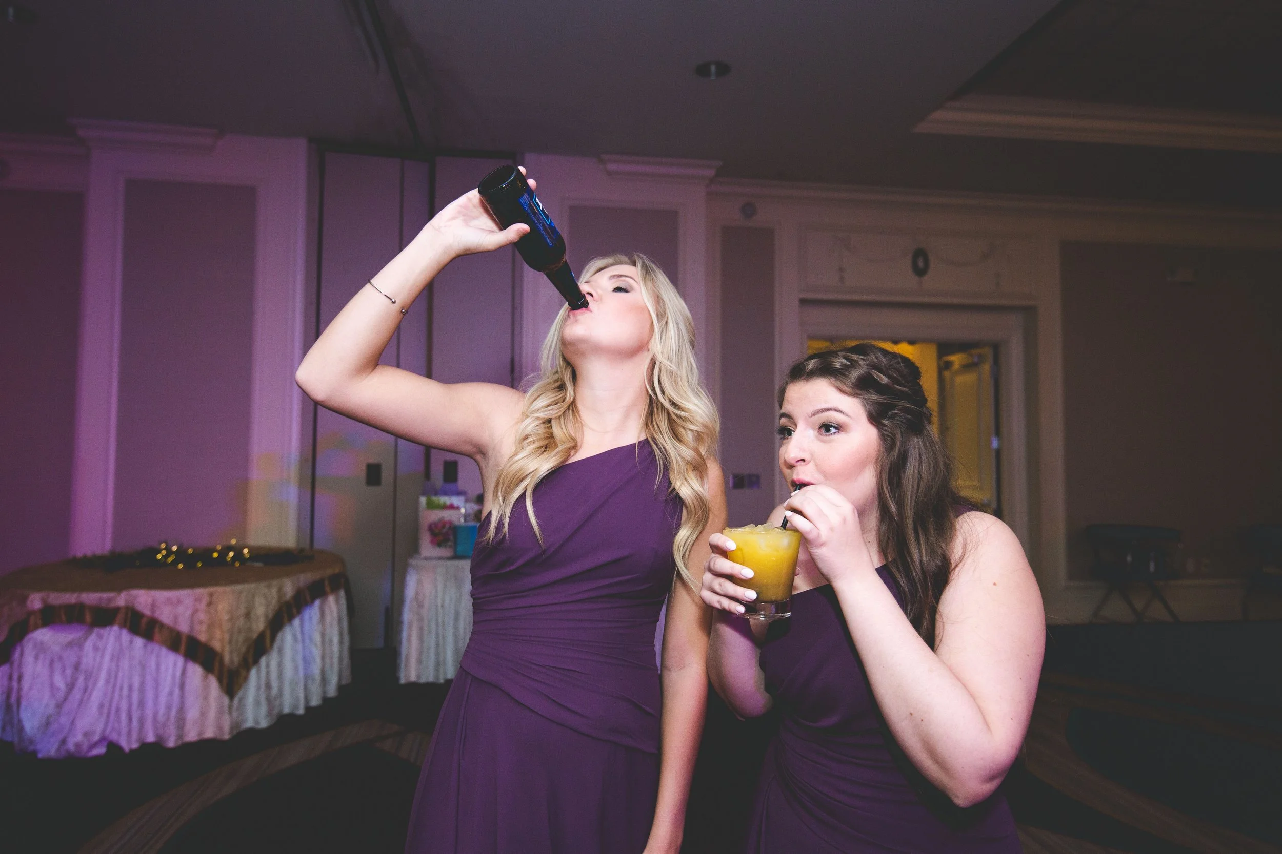 Two bridesmaids in purple dresses chug their drinks during a wedding reception with purple uplighting.