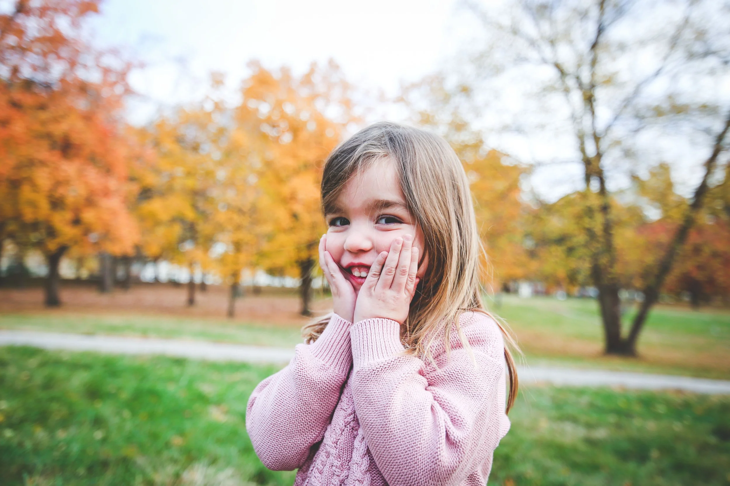 Portrait of a young girl with her cheeks in her hands smiling in front of orange and yellow fall trees.