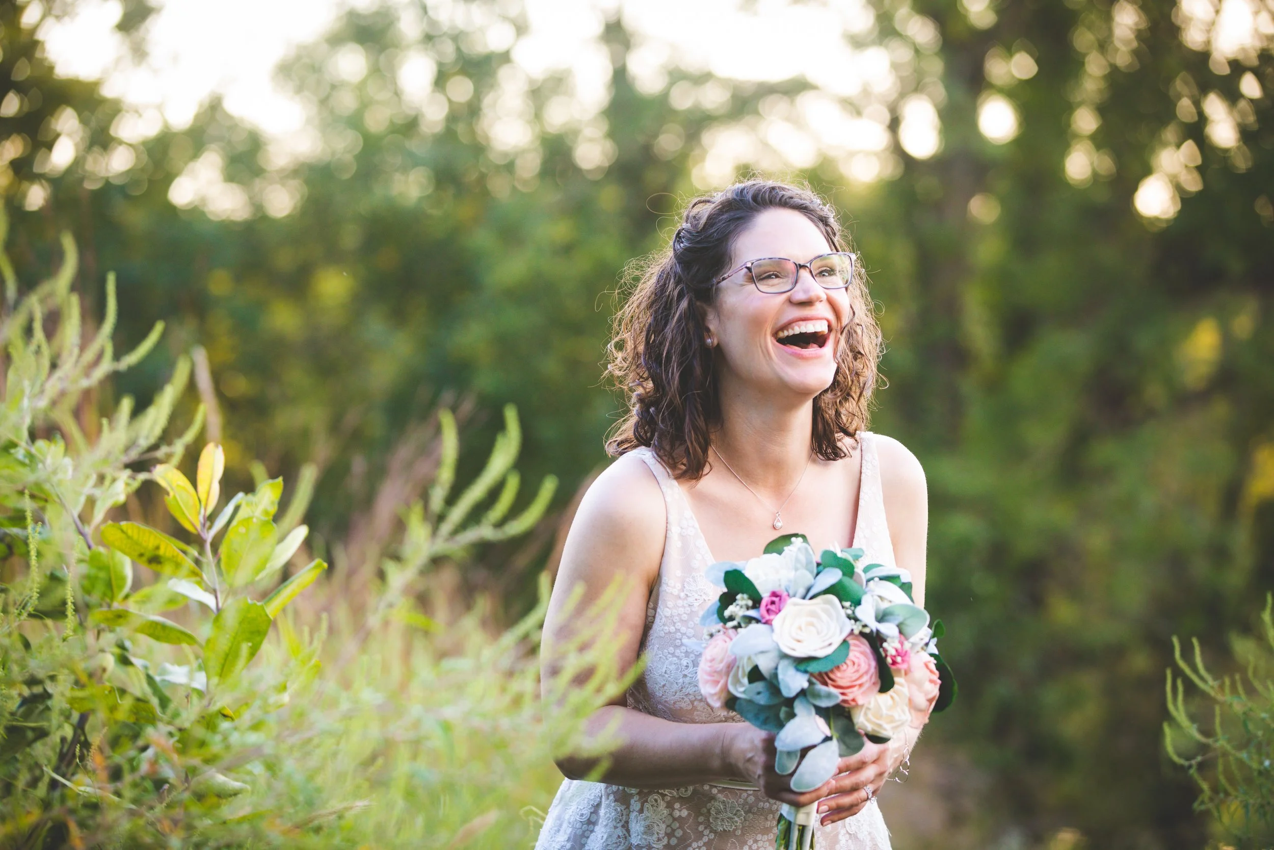 Bride Heather laughs as she holds her bouquet close in soft pink lighting among green trees during golden hour in Queeny Park in Saint Louis.
