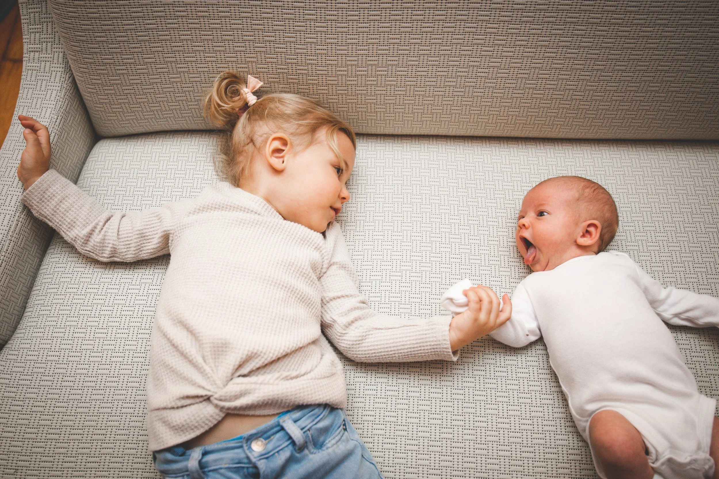 Portrait of a big sister lying with her newborn sister on the couch while holding her hand and looking at each other.