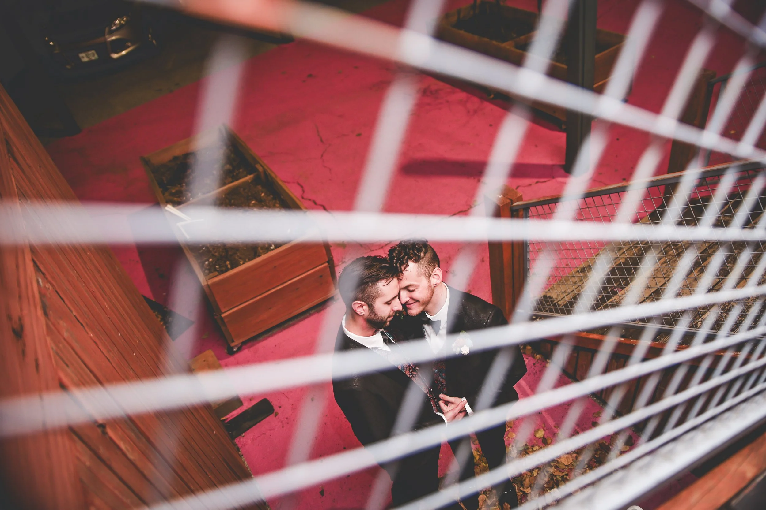 Grooms Anthony & Scott stand on a pink floor and share a private candid moment while Miha Photo captures them through a gridded metal railing.