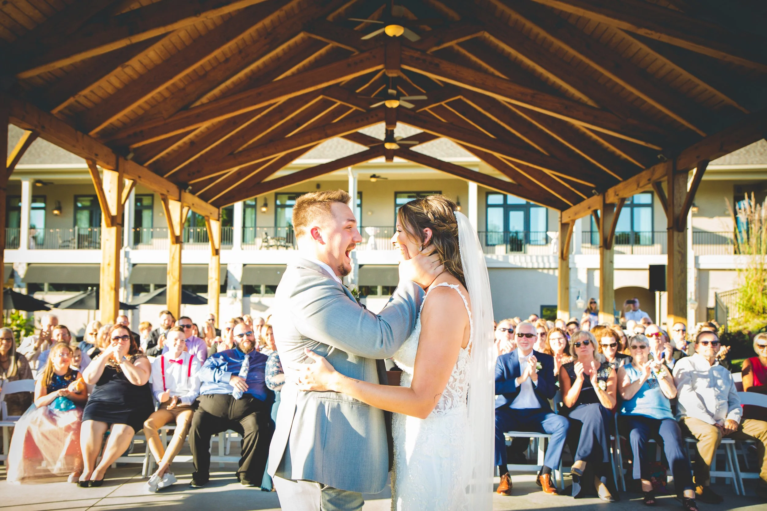 A bride and groom look so excited to share their first kiss at the altar under a wooden gazebo with clapping guests in the background.