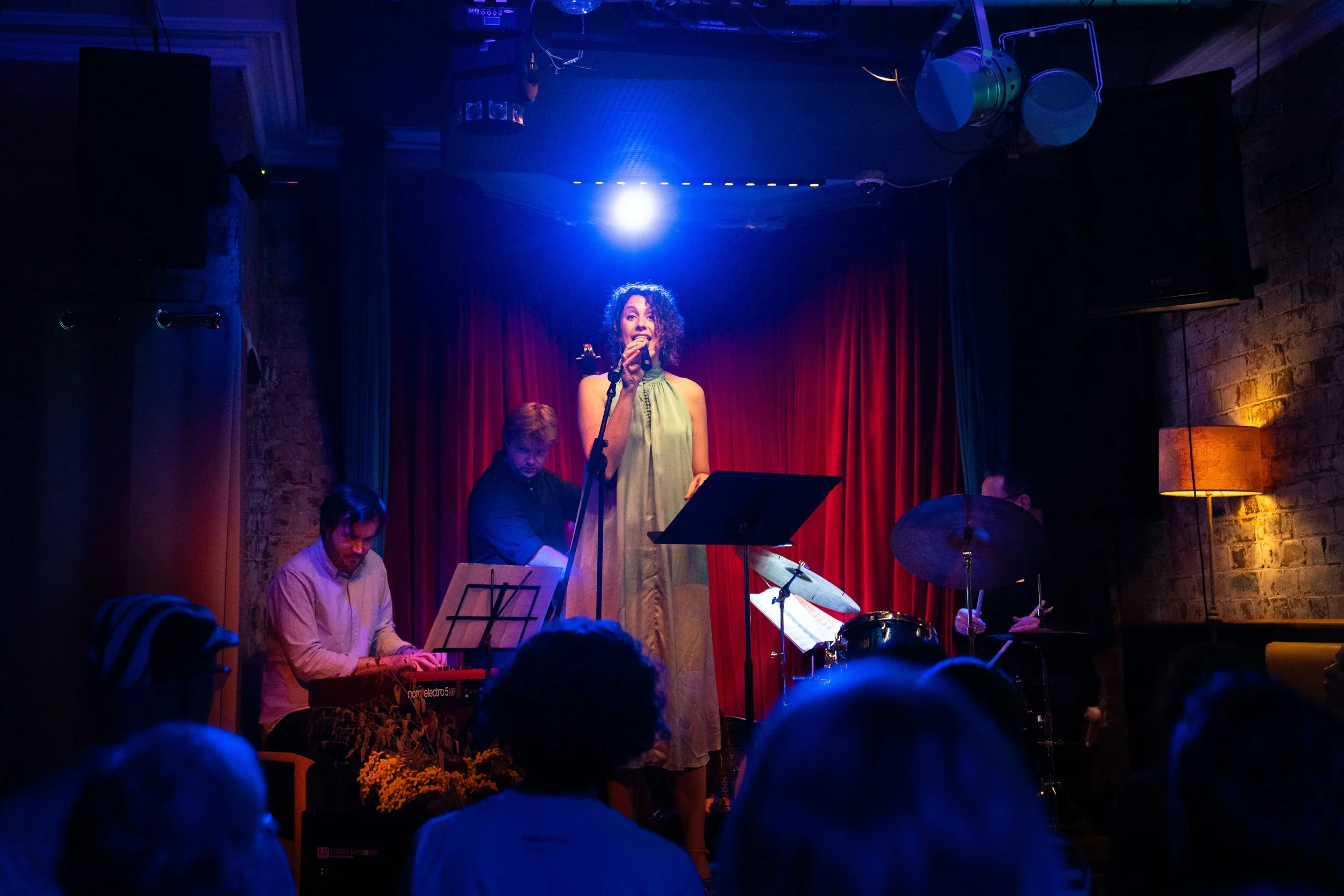A woman singing into a microphone on a stage infront of a red curatin and a jazz trio