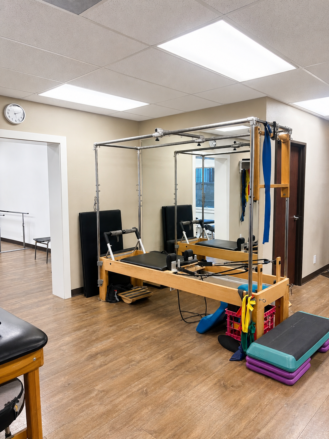 Physical therapy treatment room with Pilates reformer machine, black padded chair, exercise mats, and resistance bands inside a clinic or gym.