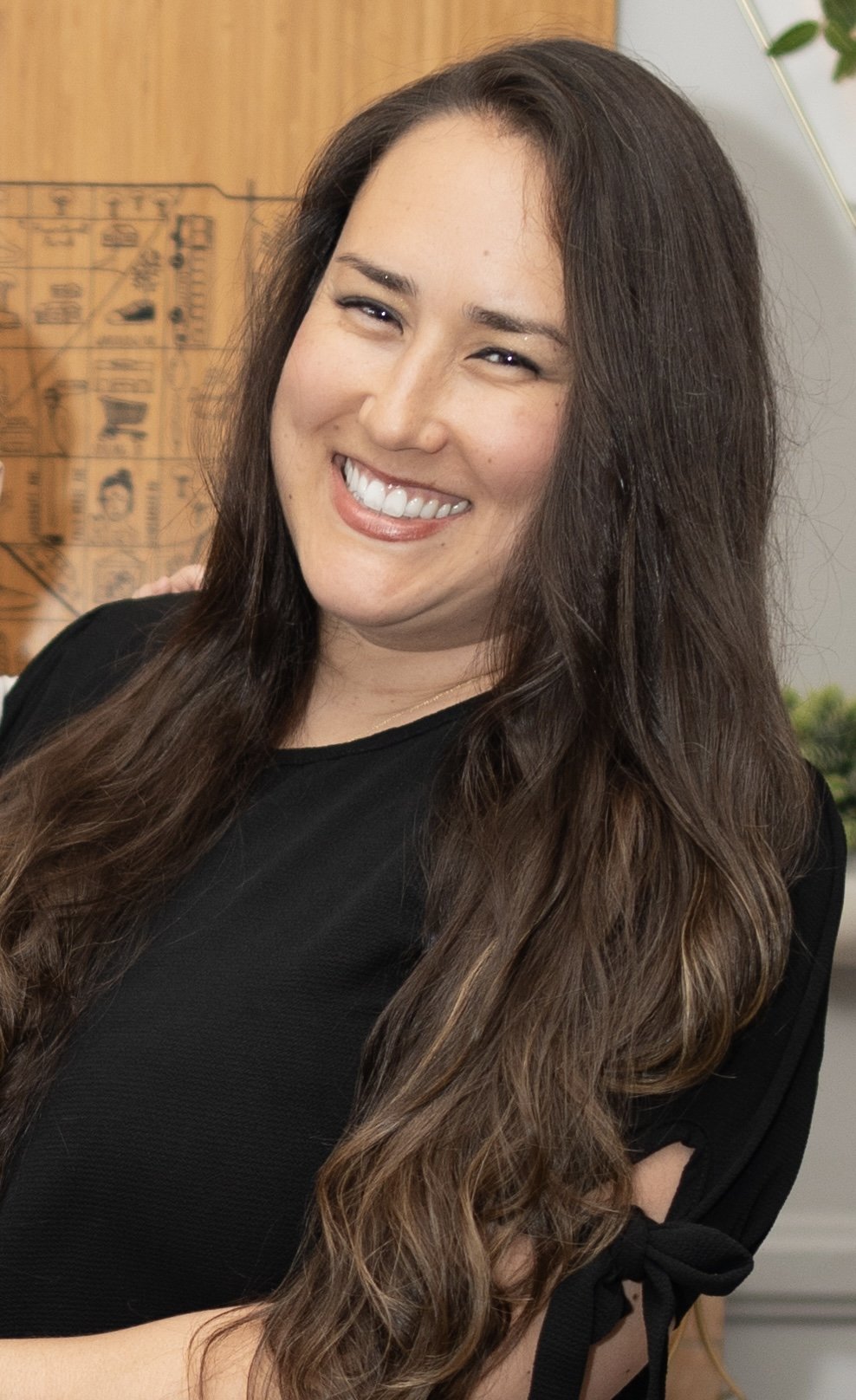 Smiling woman with long, wavy brown hair wearing a black top, standing indoors with wooden and white decor in the background.