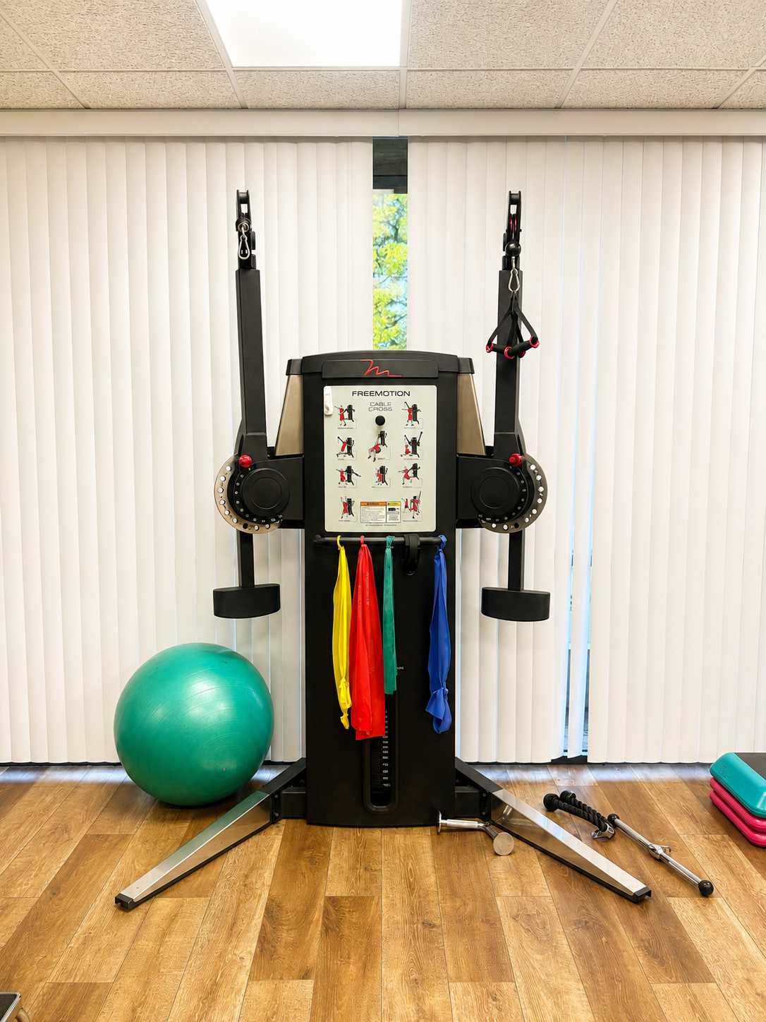 Fitness equipment in a gym room, including a wall-mounted exercise machine, a large green exercise ball, resistance bands in yellow, red, green, and blue hanging from the machine, and other exercise accessories on the floor. There are vertical blinds and a window in the background.