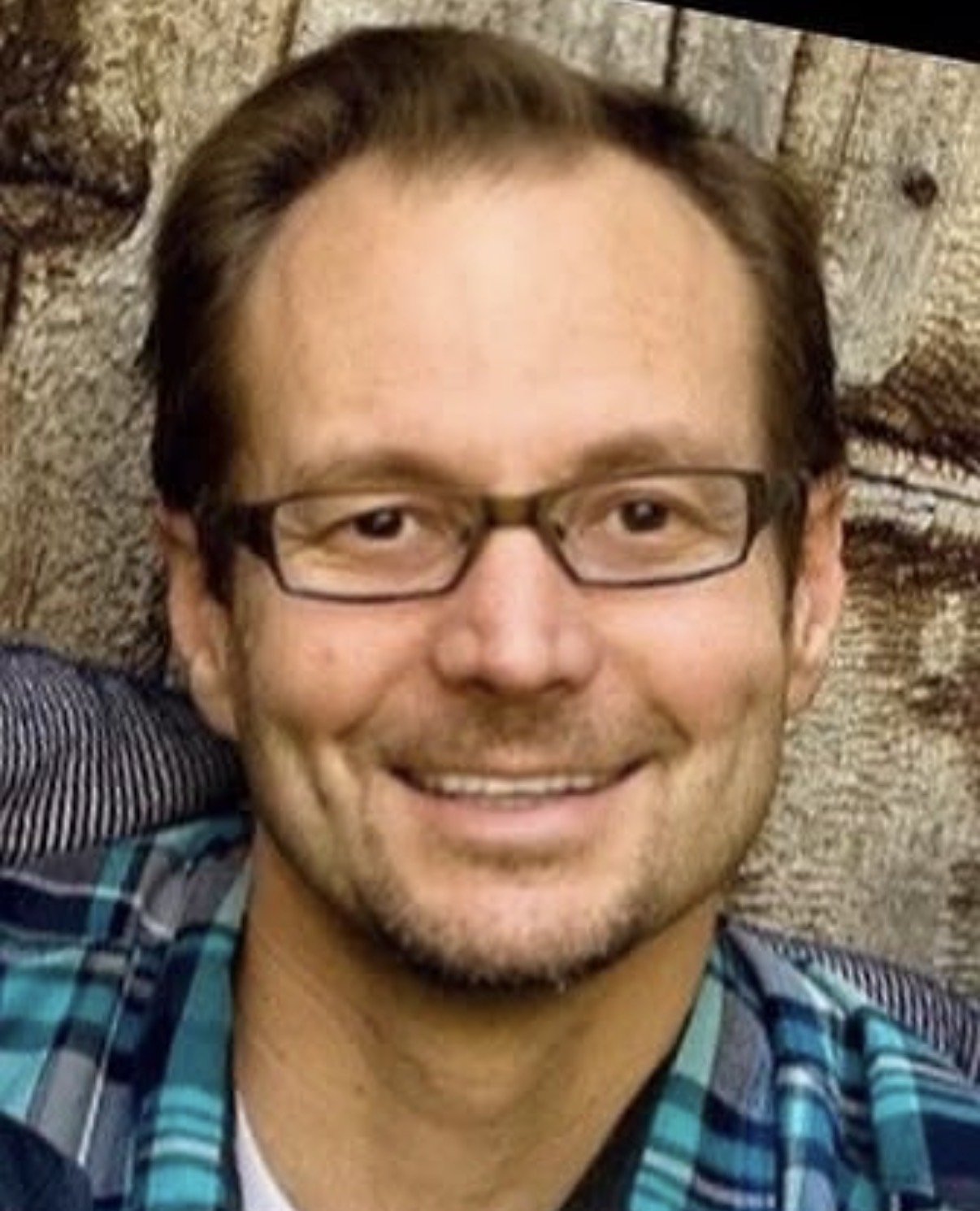 Close-up of a man smiling, wearing glasses, with brown hair, a beard, and a blue plaid shirt, lying on a textured, brown and gray surface.