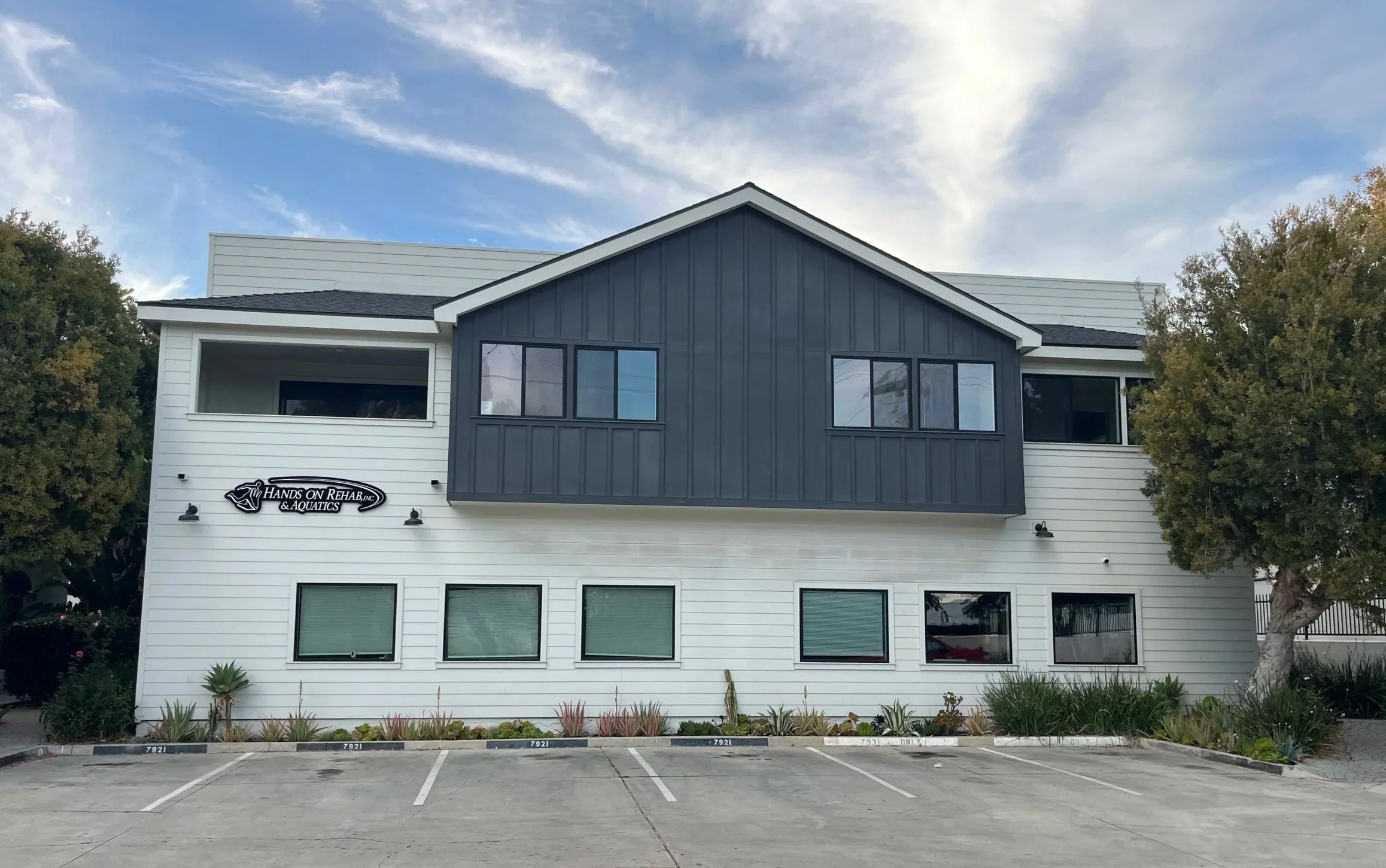 A modern two-story building with white and dark gray exterior siding, multiple large windows, and a sign for 'Hands on Rehab & Aquatics' on the lower left side. There are trees on each side of the building and a parking lot in the foreground.