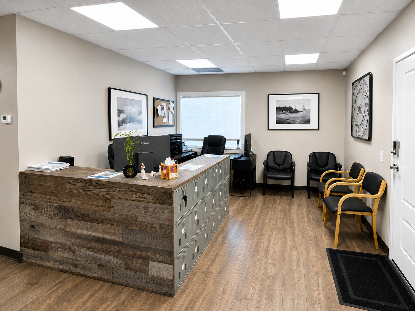 An office reception area with a wooden front desk, black chairs, a computer, framed black and white photos on the beige walls, and a window with blinds.