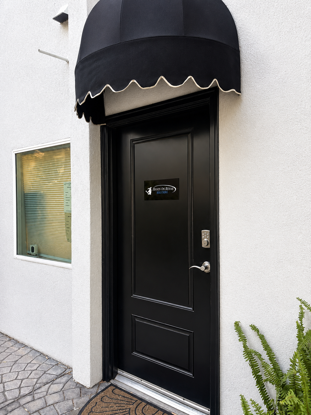 Black door with a black awning above it, leading into a business named 'Hands on Rehab.' A window with closed blinds is to the left, and green plants are to the right. The door has a keypad lock.