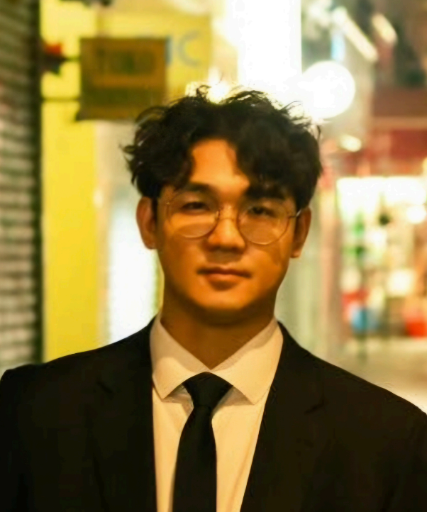 A young man with dark curly hair and glasses, dressed in a black suit, white shirt, and black tie, standing outdoors at night in an urban area with blurred colorful lights in the background.
