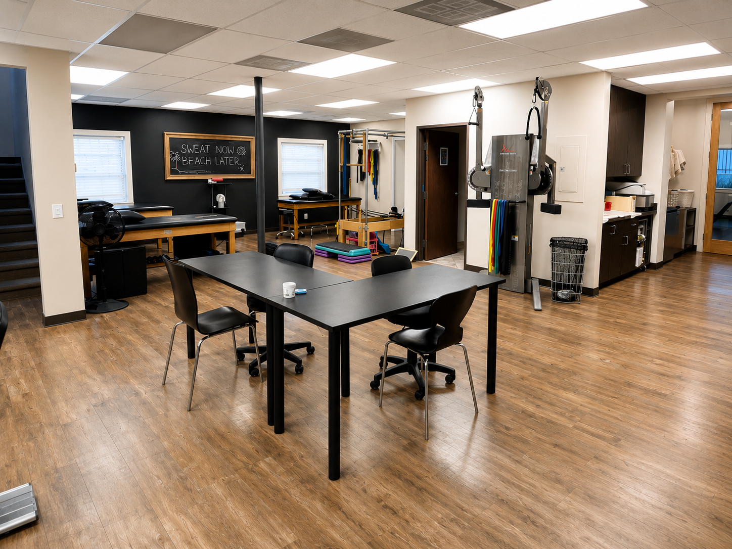 Rehab or physical therapy room with exercise equipment, black chalkboard with handwritten message, table with chairs, and wood flooring.
