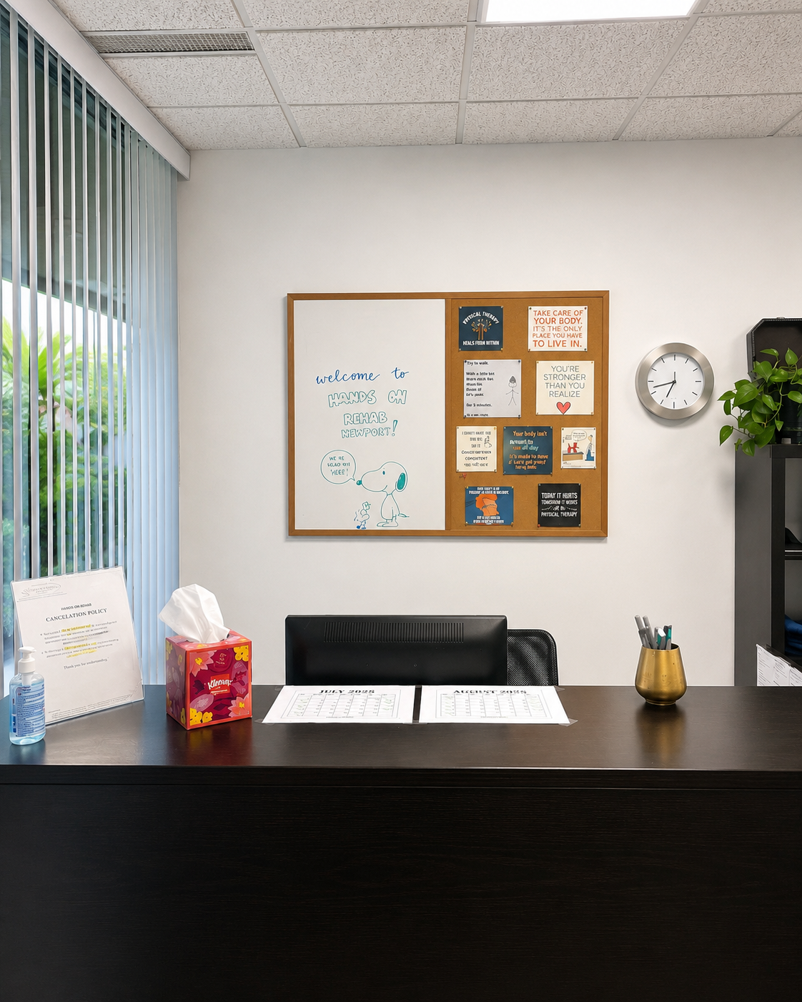 Office reception desk with a whiteboard and bulletin board on the wall. Items on the desk include a tissue box, hand sanitizer, and a pen holder. There is a clock and a plant on a shelf. The office has vertical window blinds and a white ceiling with ceiling tiles.