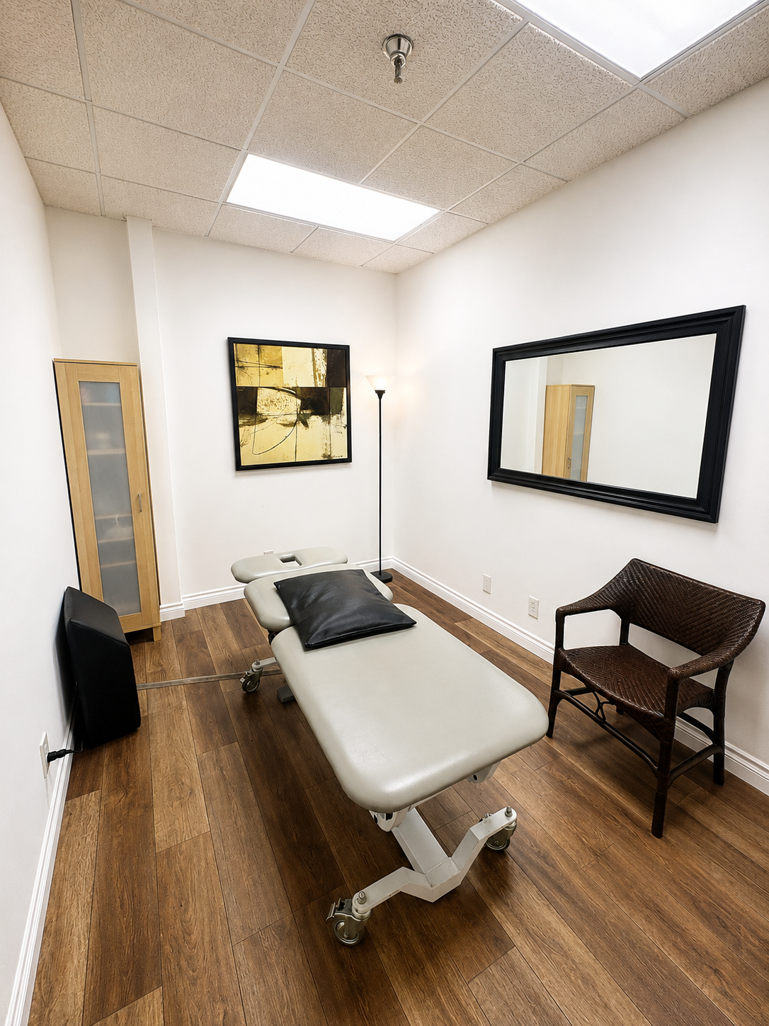 A physical therapy examination room with a white adjustable treatment table, a black pillow, a black framed mirror on the wall, a wooden cabinet, a brown wicker chair, a tall floor lamp, and a piece of abstract artwork on the wall.