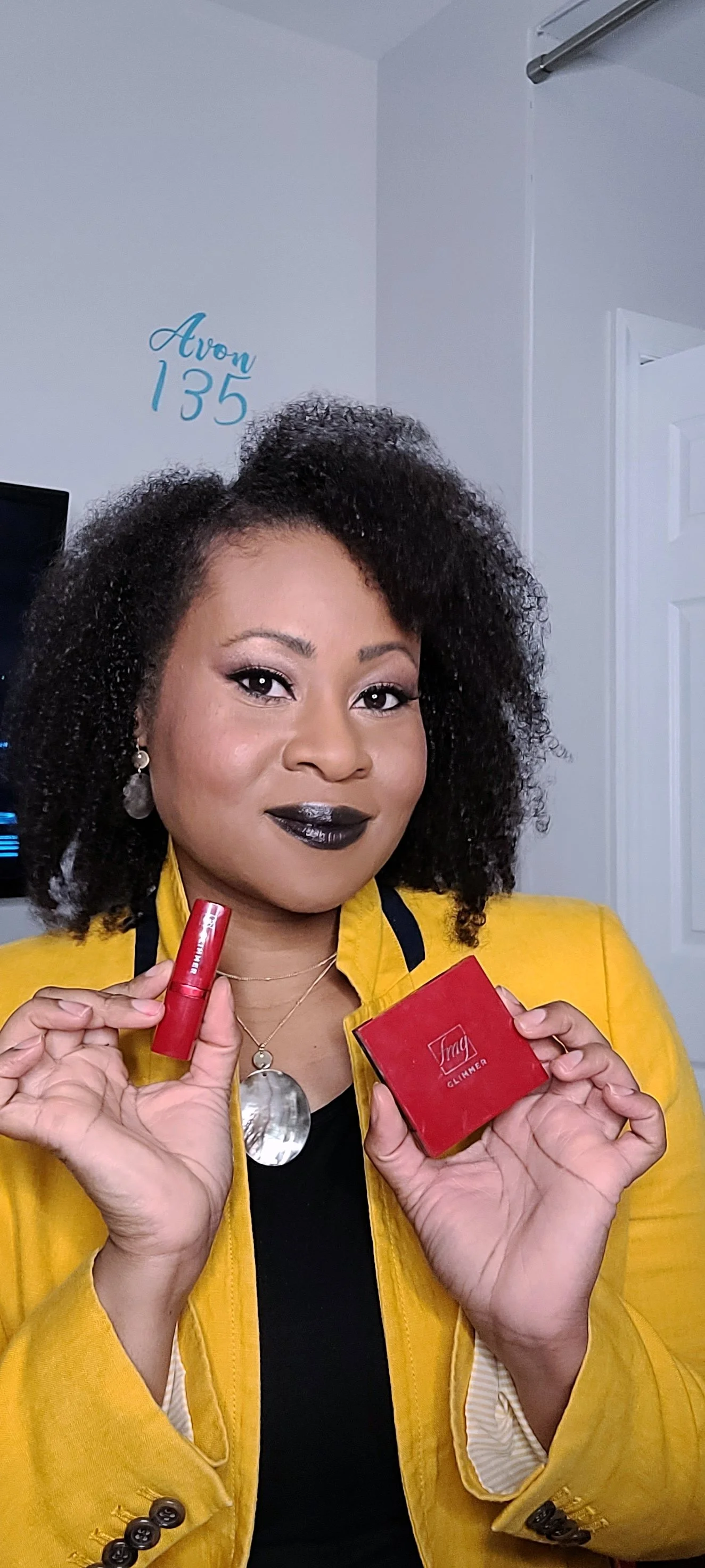 black woman with natural hair, black lipstick, and yellow j. crew blazer, holding Avon lipstick and eyeshadow products