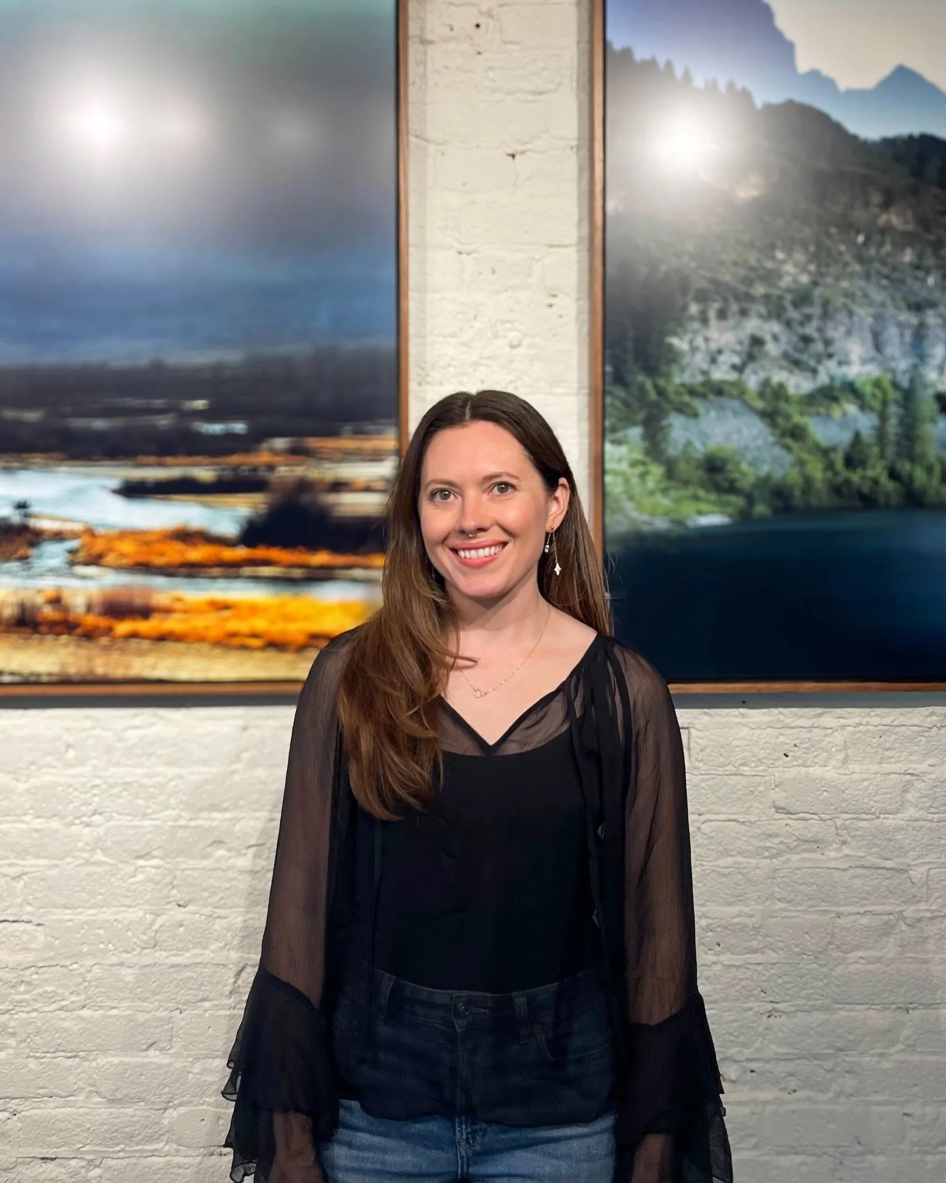 A woman with long brown hair, smiling, wearing a black sheer long-sleeve top and earrings, standing in front of a white brick wall with two landscape paintings, one showing a coastal scene with water and sky, and the other a mountainous terrain with trees and rocks.