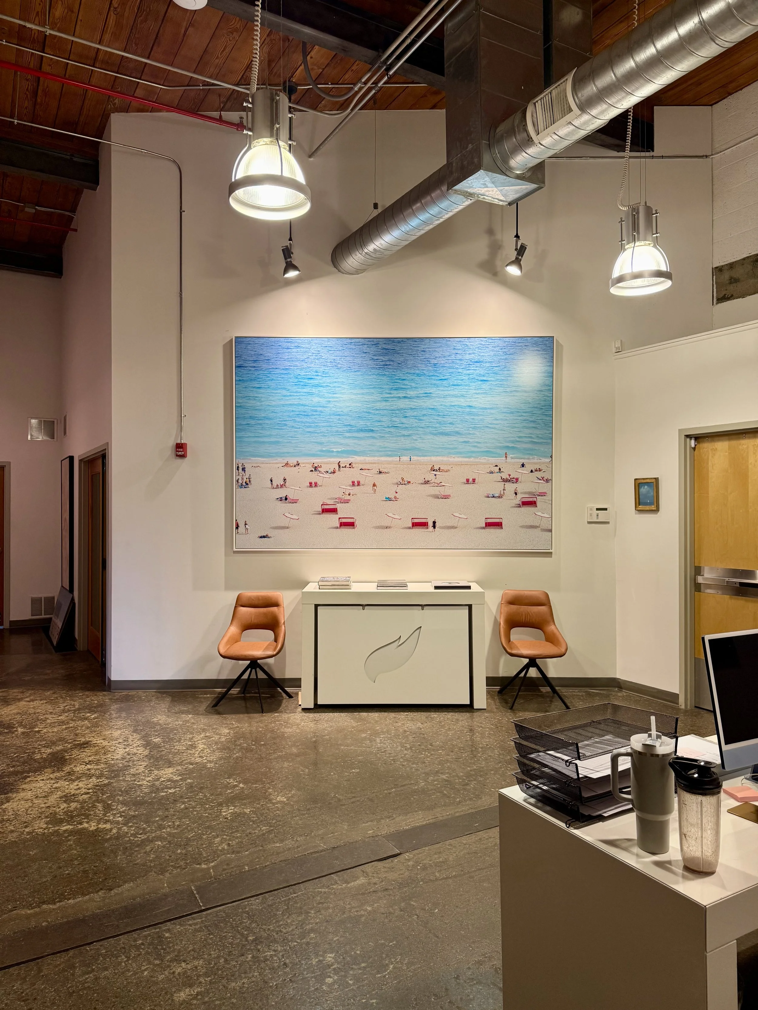 An interior office space with a large beach scene painting on the wall, two orange chairs, a white counter with a leaf logo, and various office supplies in the foreground.