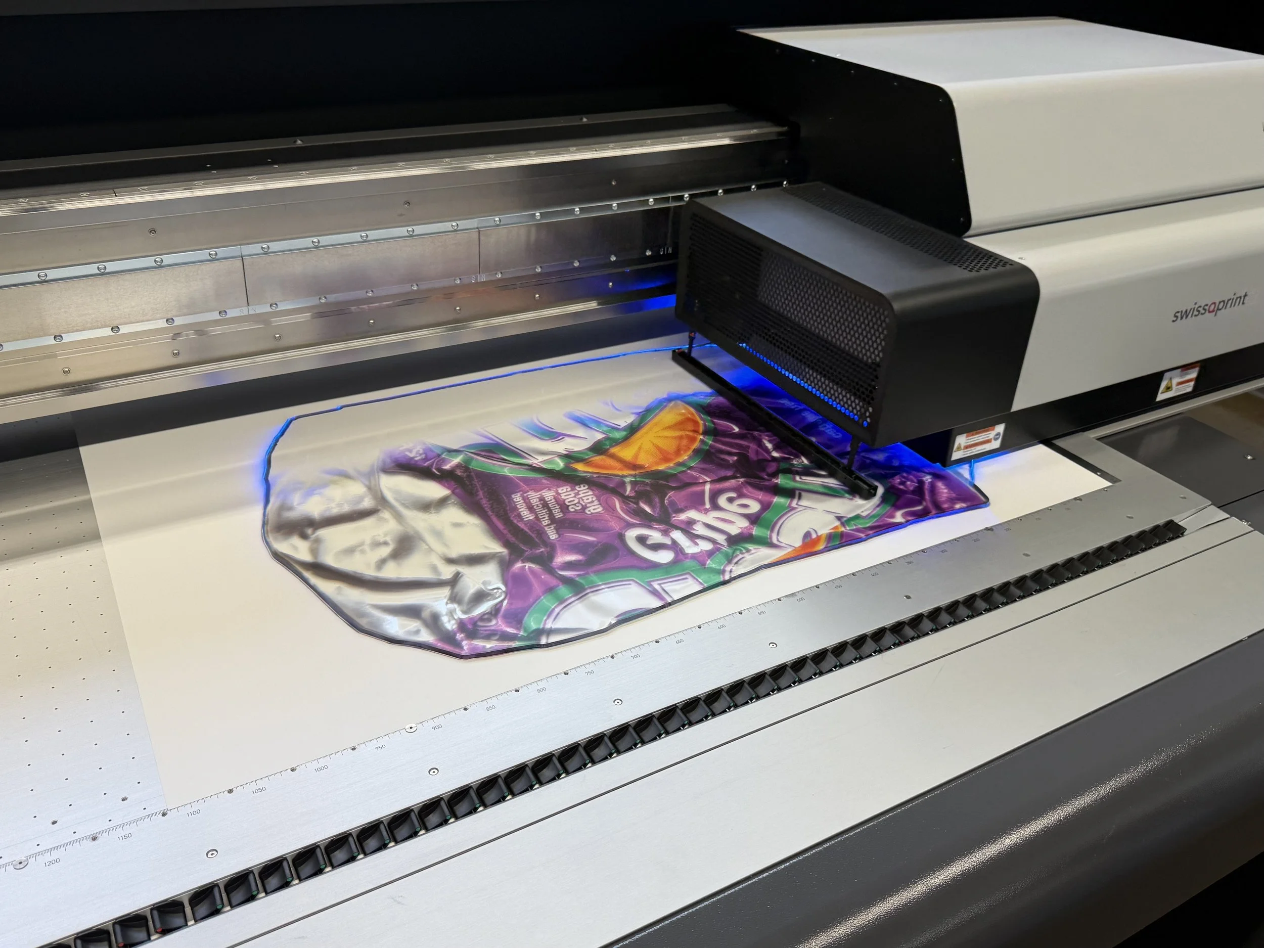 A large flatbed printer printing a colorful image on a fabric or similar material. The printer has a metal frame with measurement markings, and the image being printed is vibrant with purple, orange, and green colors.