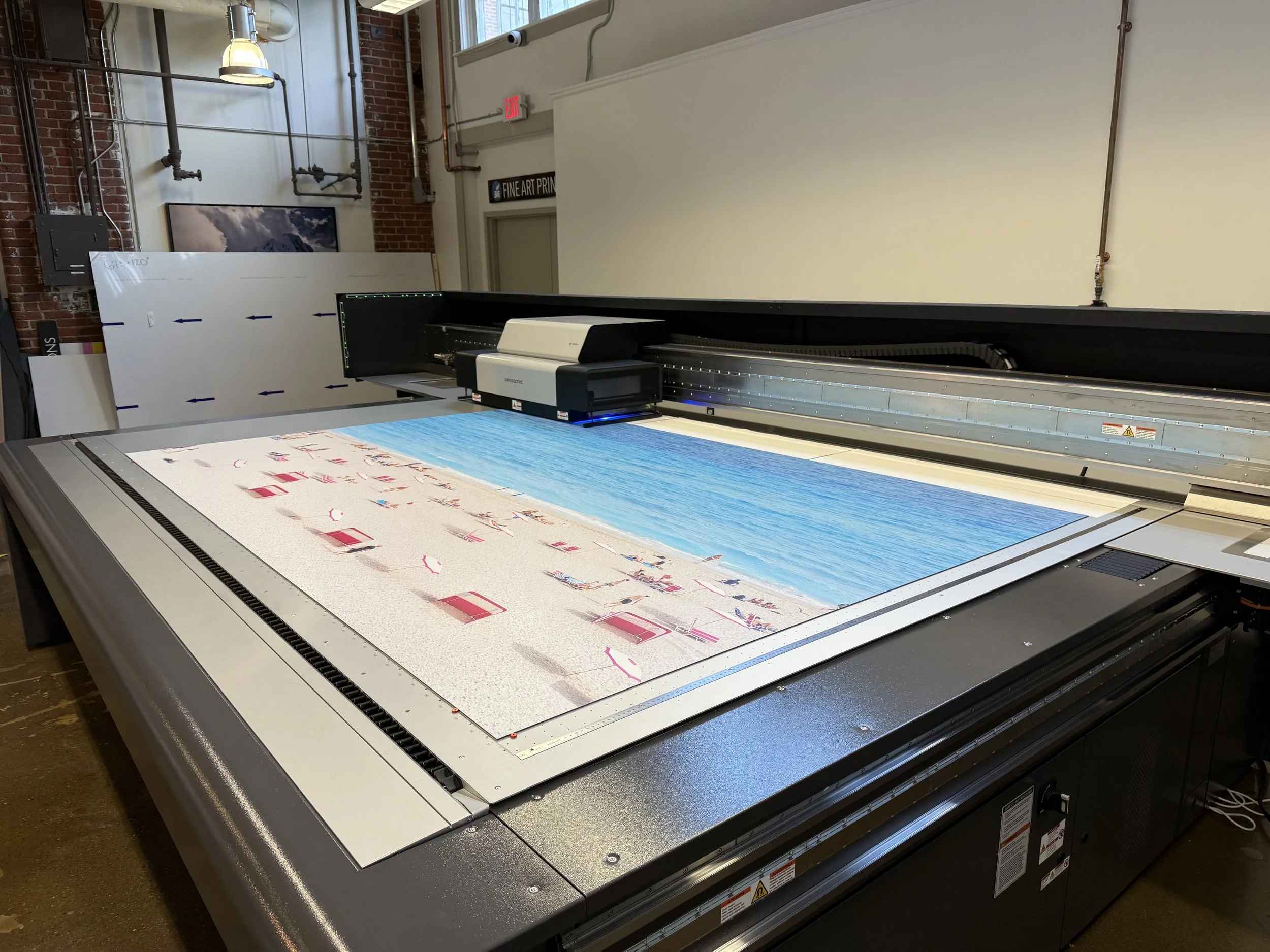 Large flatbed printer printing a colorful image of a blue sky and pink abstract shapes. The printer is in an industrial-style room with brick walls and visible piping.