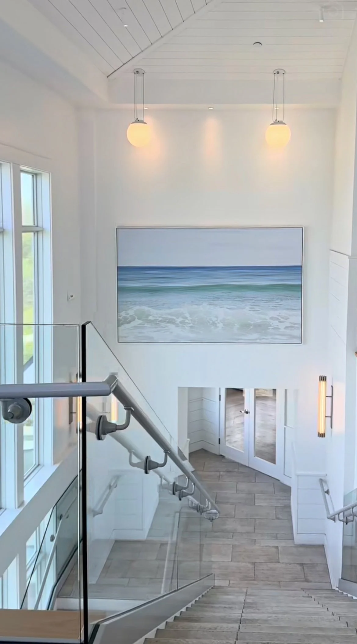 Interior view of a modern beach house with a large painting of the ocean on the wall, stairs, and large windows letting in natural light.