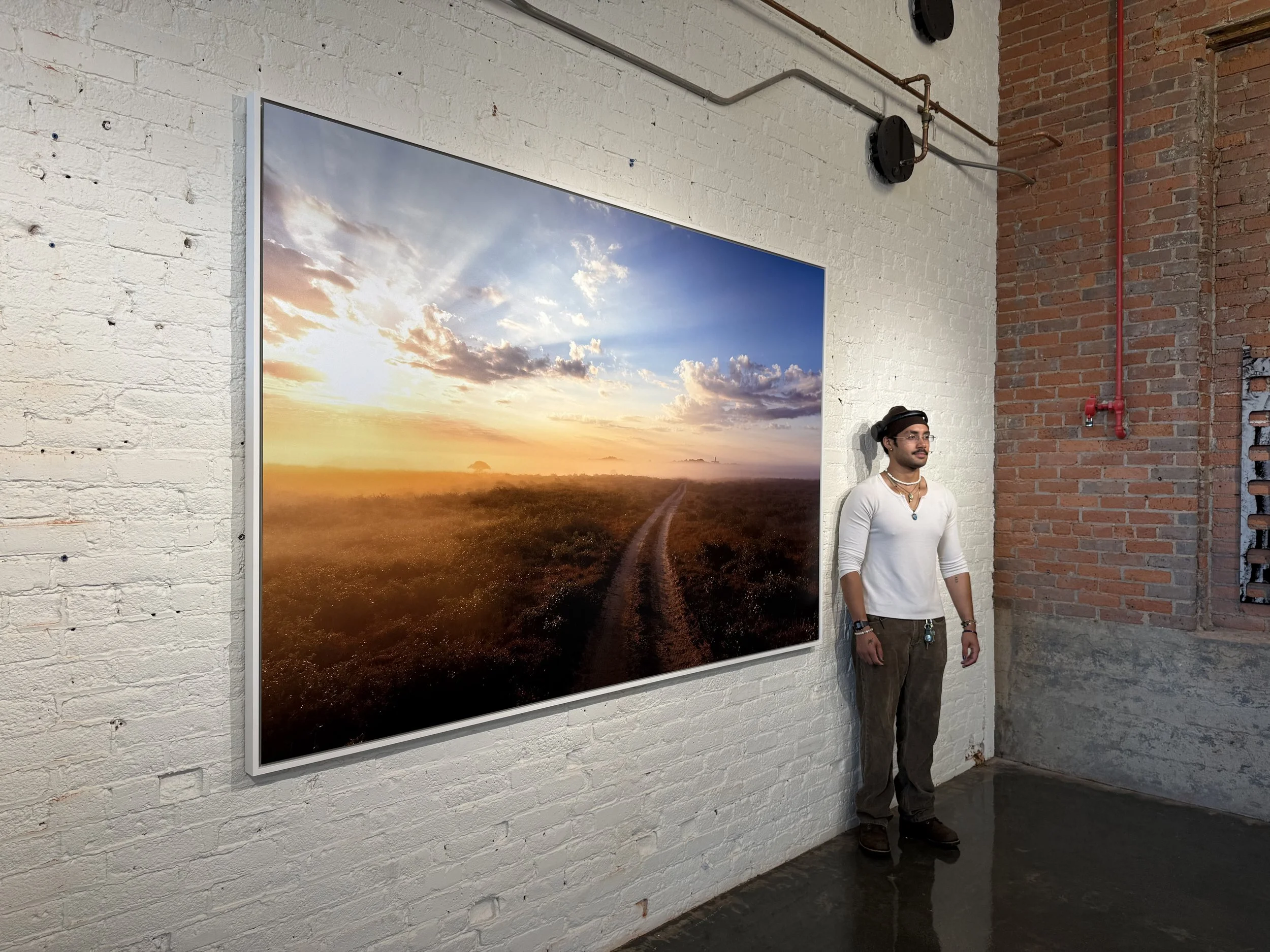 A man with glasses, wearing a white long-sleeve shirt, a necklace, and a hat, stands next to a framed landscape photograph of a sunset over a field, hanging on a white brick wall in an art gallery.
