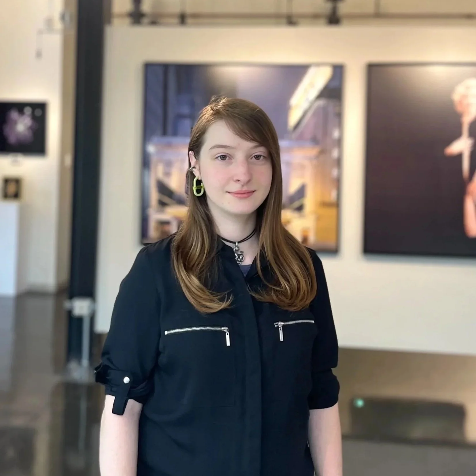 A young woman with long brown hair, wearing a black jacket with silver zippers, standing in an art gallery with framed artwork behind her.