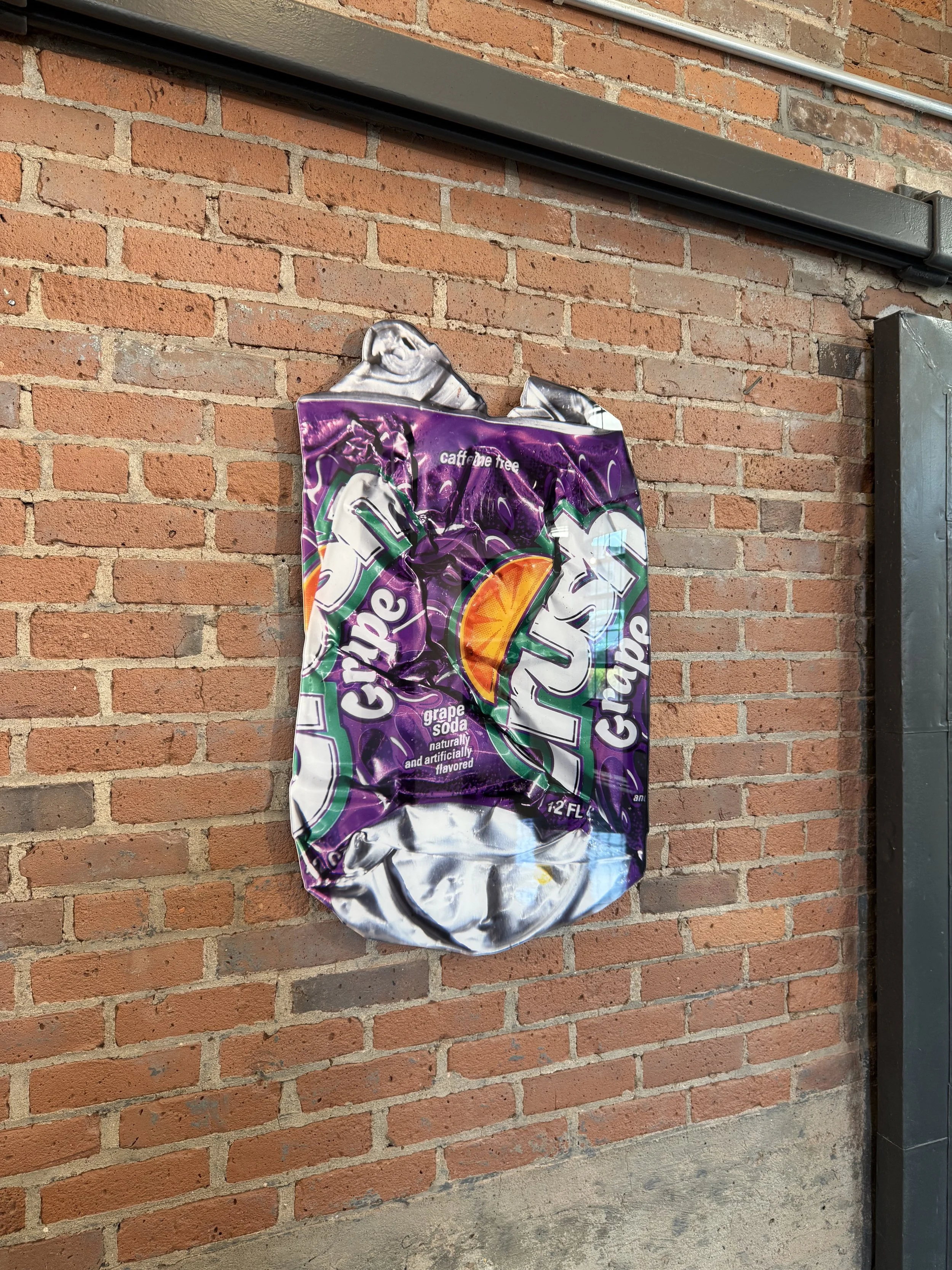 A crumpled grape soda bag hanging on a brick wall, near a metal pipe and an electrical box.