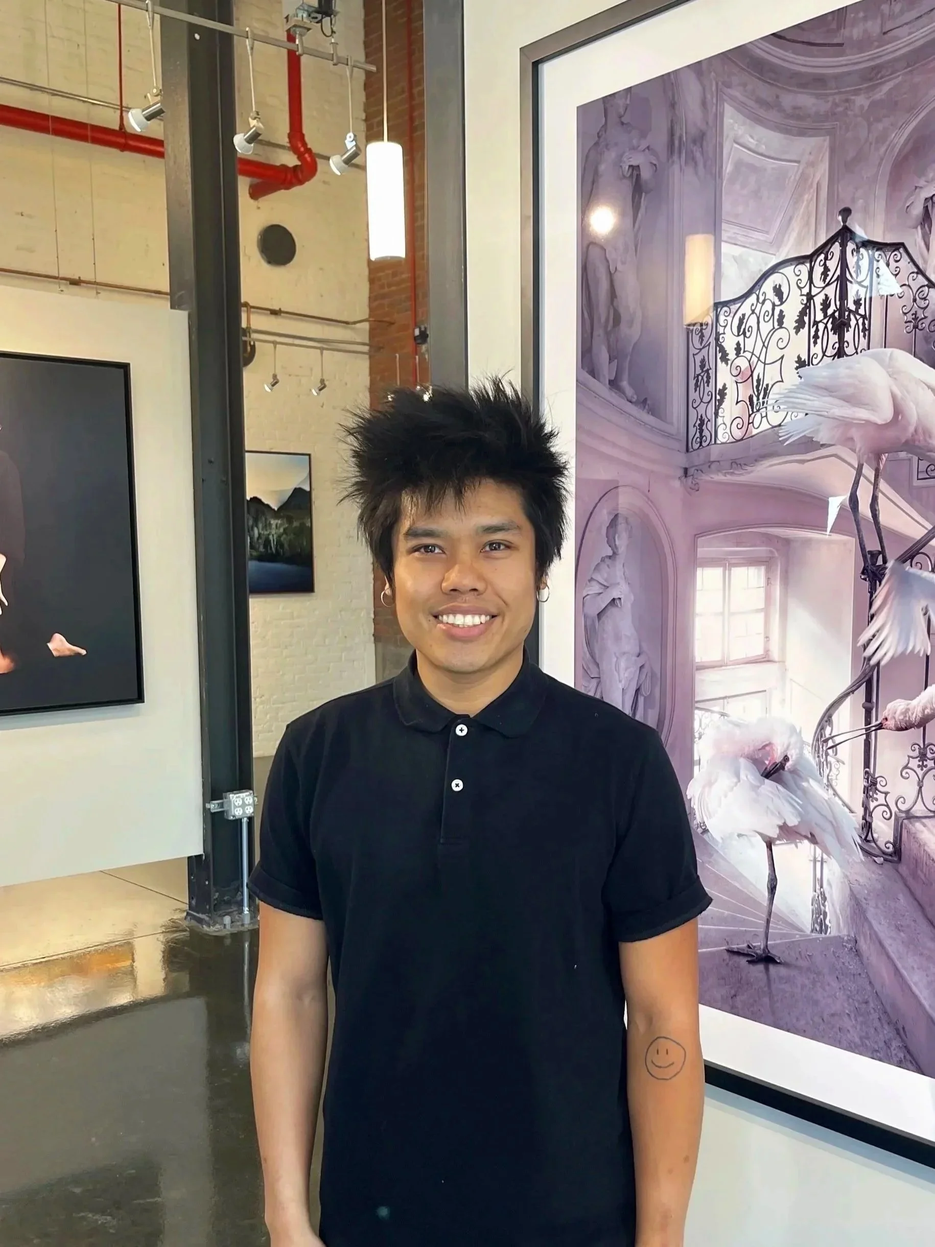 A young man with spiky black hair and earrings, smiling, standing in front of artwork depicting flamingos and classical sculptures.