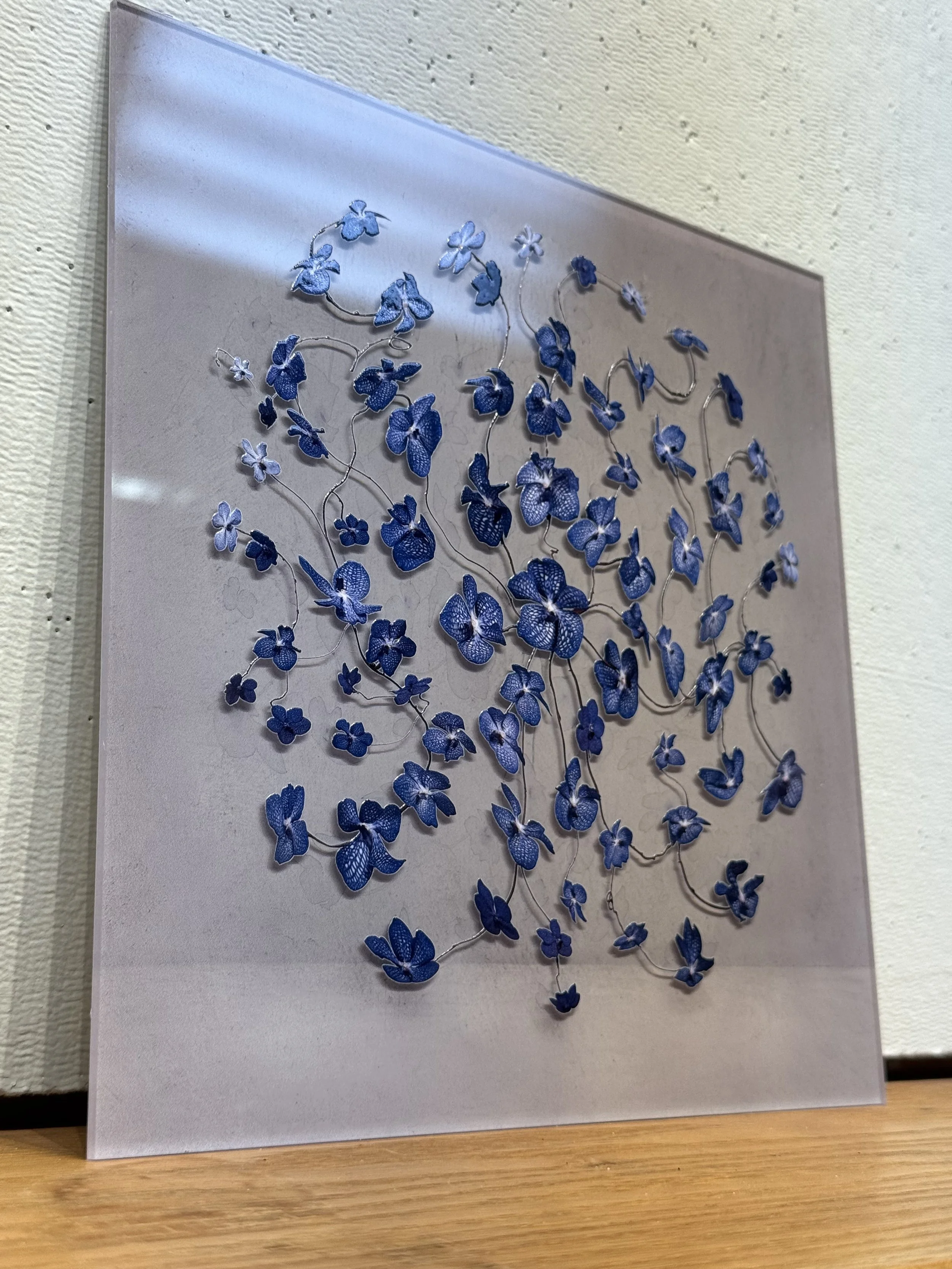 A framed artwork featuring a 3D design of numerous small blue and purple flowers connected by silver wires, mounted on a gray background and displayed on a wall.