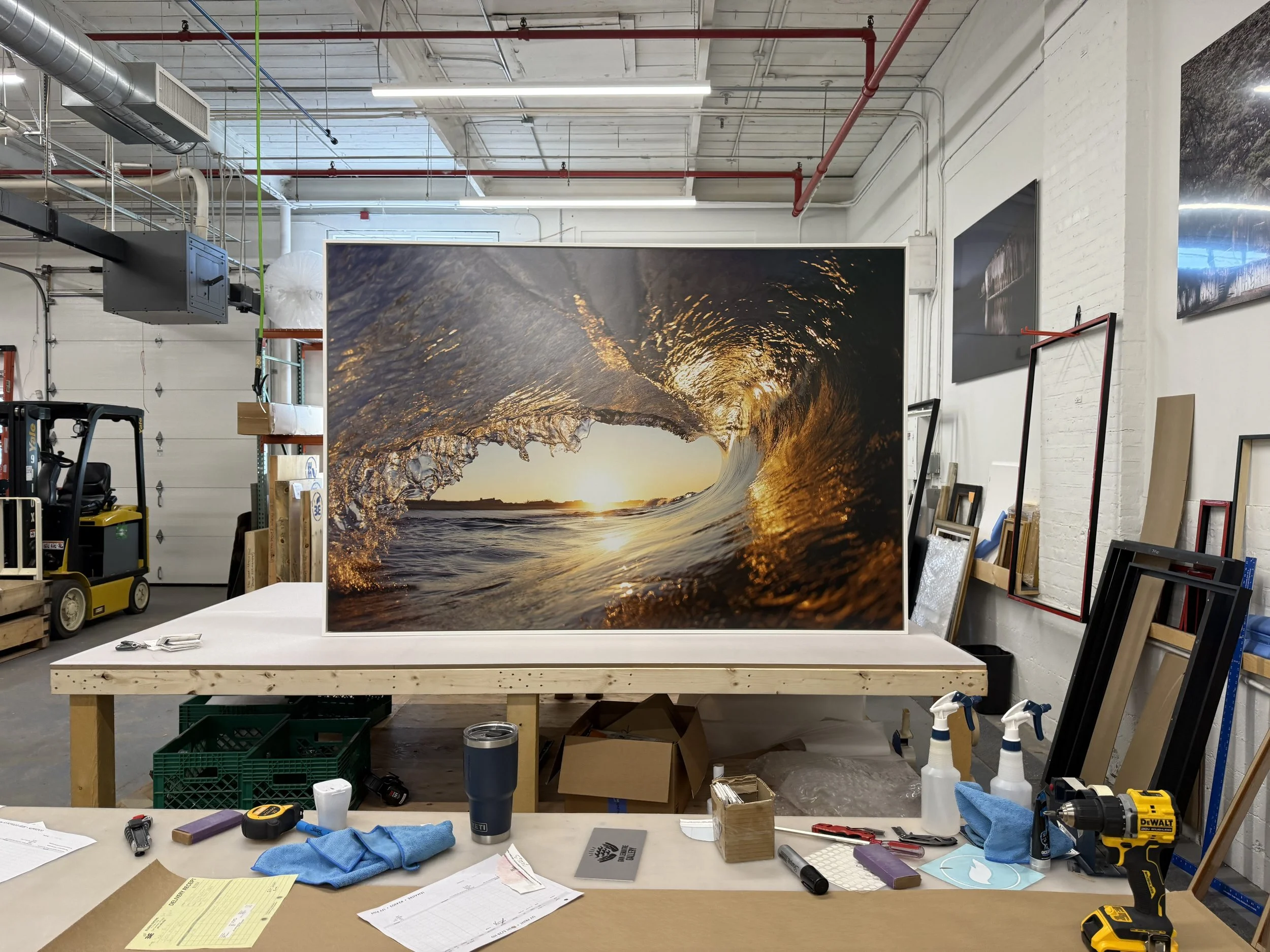 An art gallery workspace with a large photograph of a wave at sunset in the background. The table in the foreground has tools, papers, and supplies for artwork or framing, with a white brick wall and artwork on display.