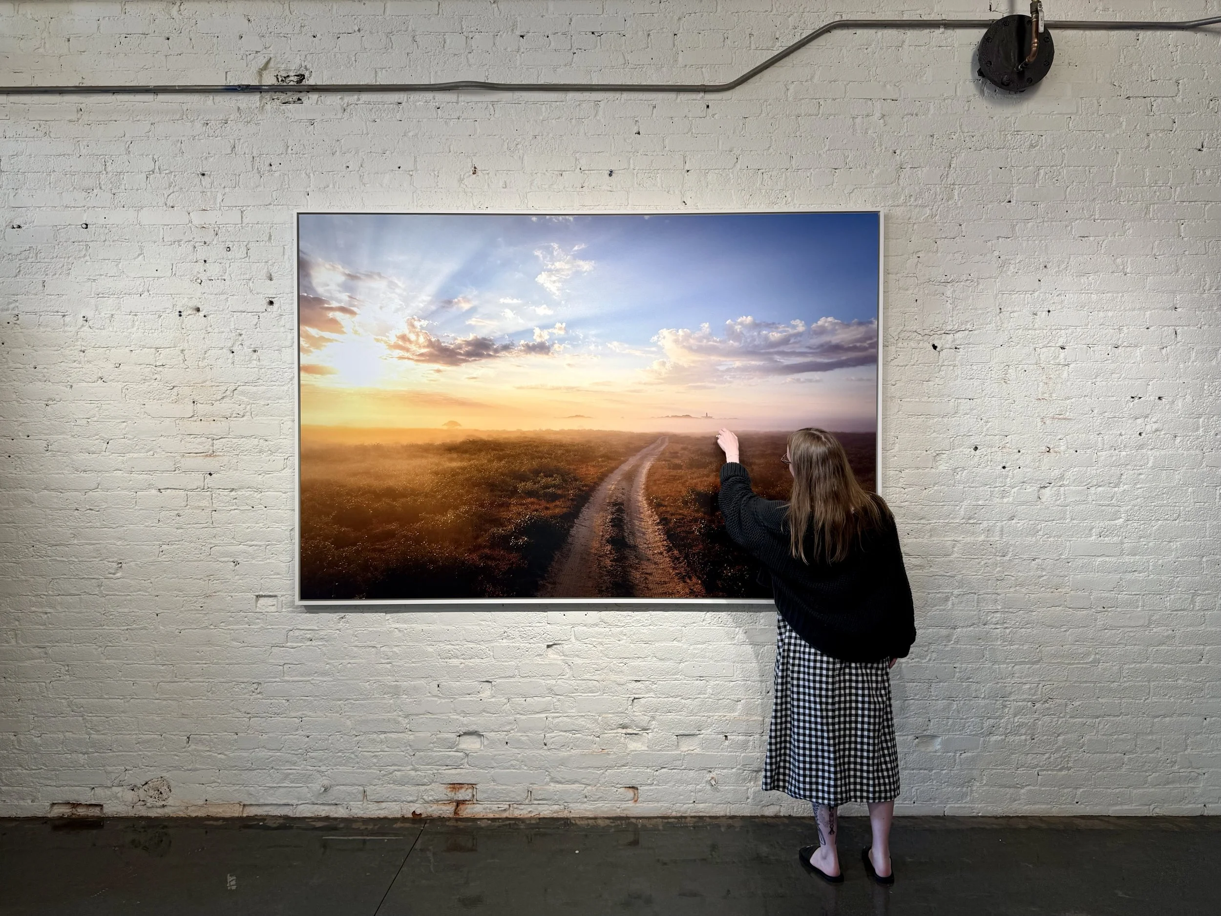 A person with long hair, wearing a black sweater and a checkered skirt, standing in front of a large artwork depicting a sunset over a dirt path in an open field, with their hand touching or pointing at the art.