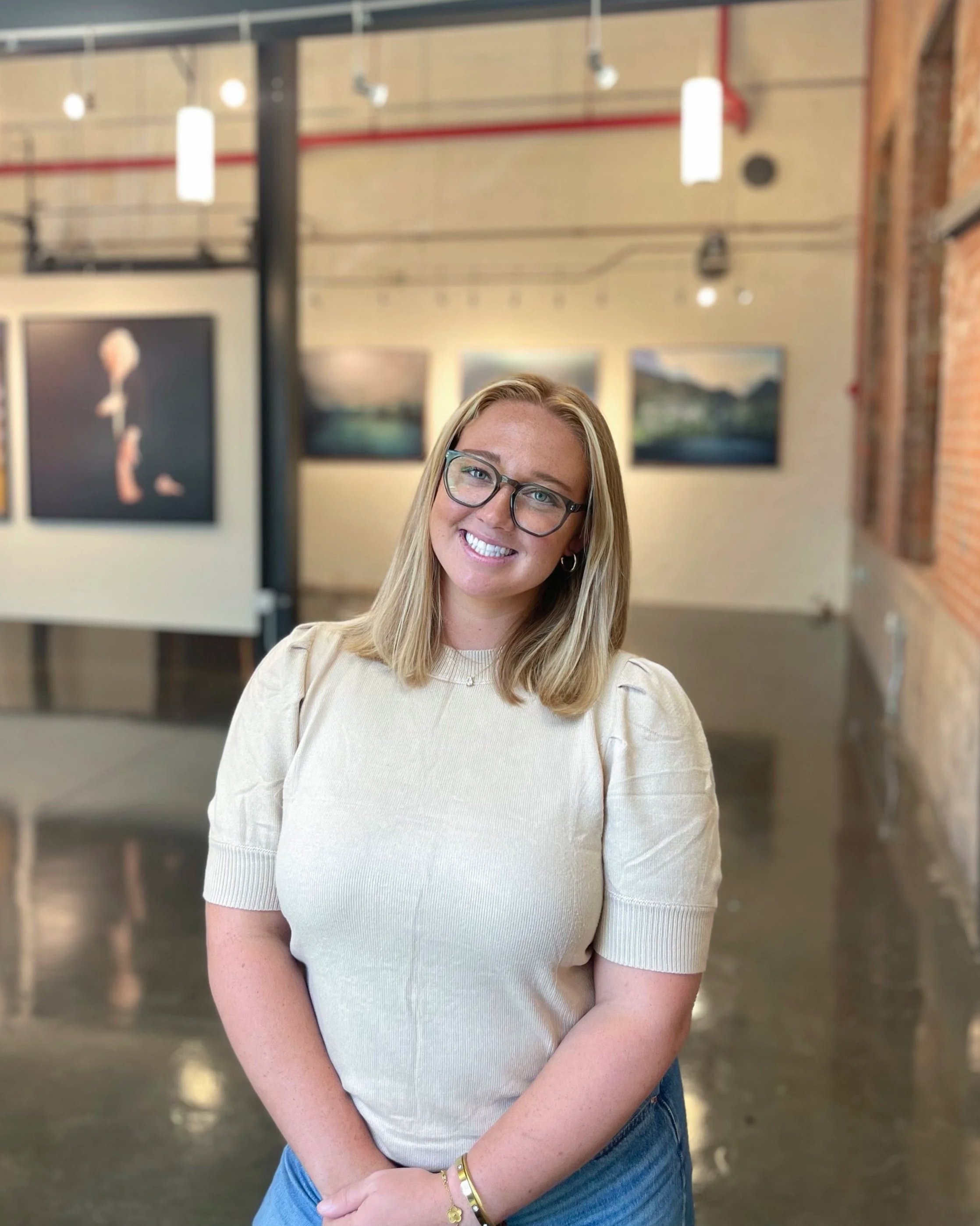 A smiling woman with blonde hair and glasses, wearing a light-colored sweater, standing in an art gallery with paintings on the wall behind her.