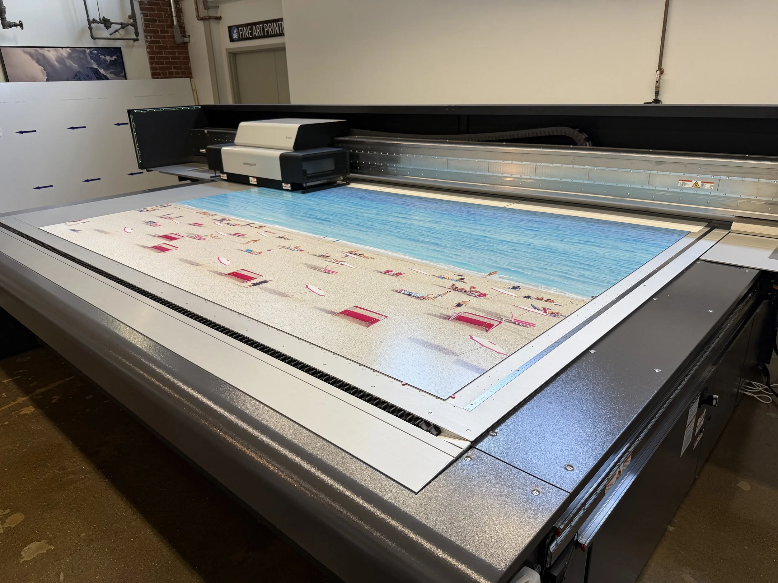A large flatbed scanner or printer in an industrial or office setting, with a printed beach scene photograph on the scanning surface. The scanner has a black and white body with control panels and a cover open, showing the printed image.