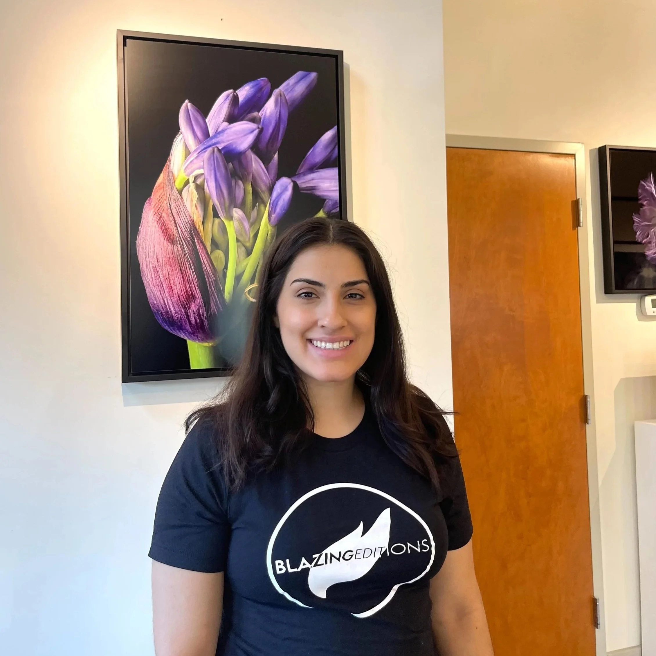 A woman with long dark hair smiling at the camera, wearing a black T-shirt with white text that says 'Blazingditions'. She is indoors with a white wall and a wooden door in the background, and there is a framed digital art print of purple flowers on the wall.