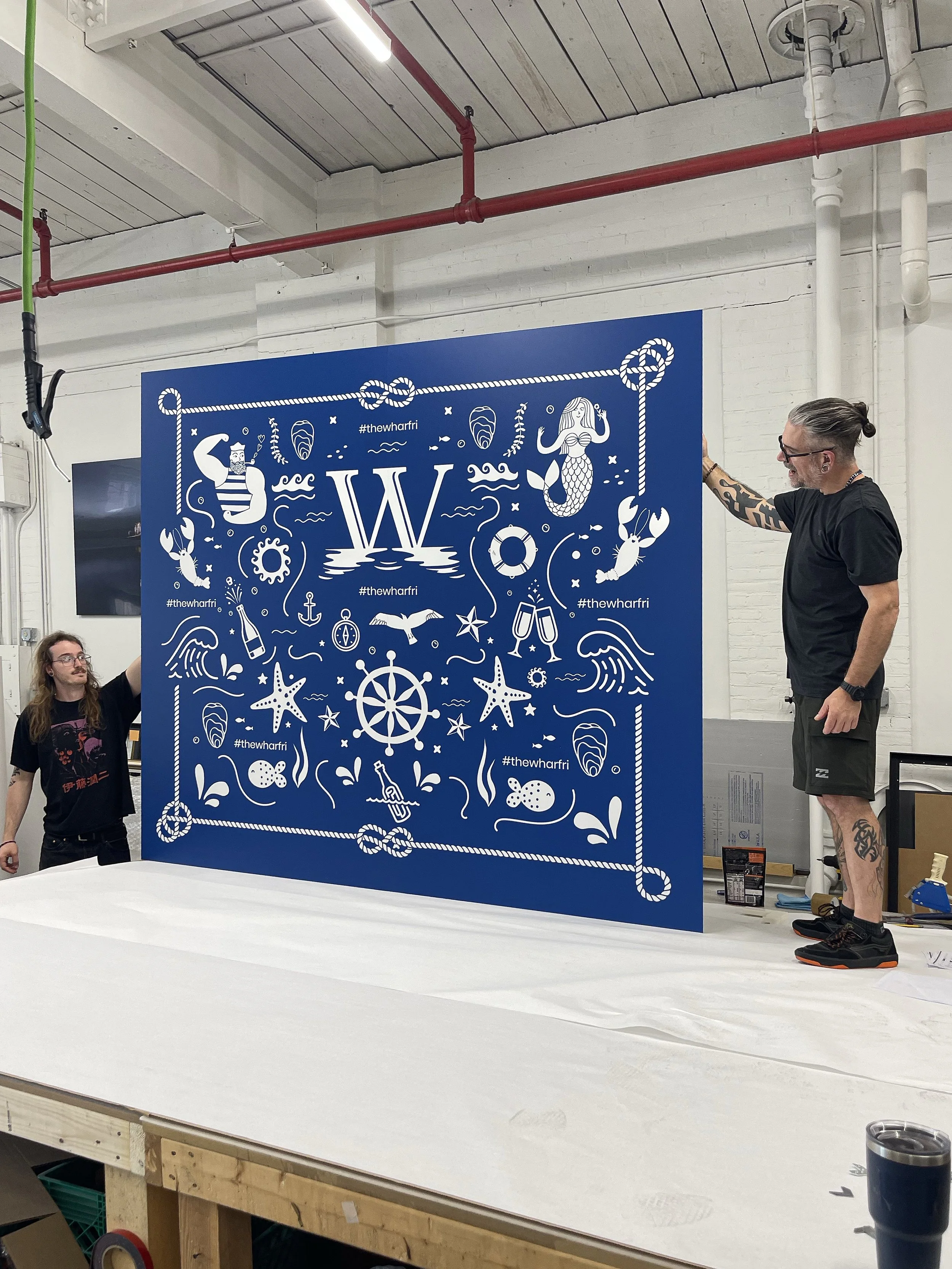 People working on a large blue wall with marine-themed illustrations, including mermaids, seahorses, starfish, and ships' wheels, in a workshop or studio.