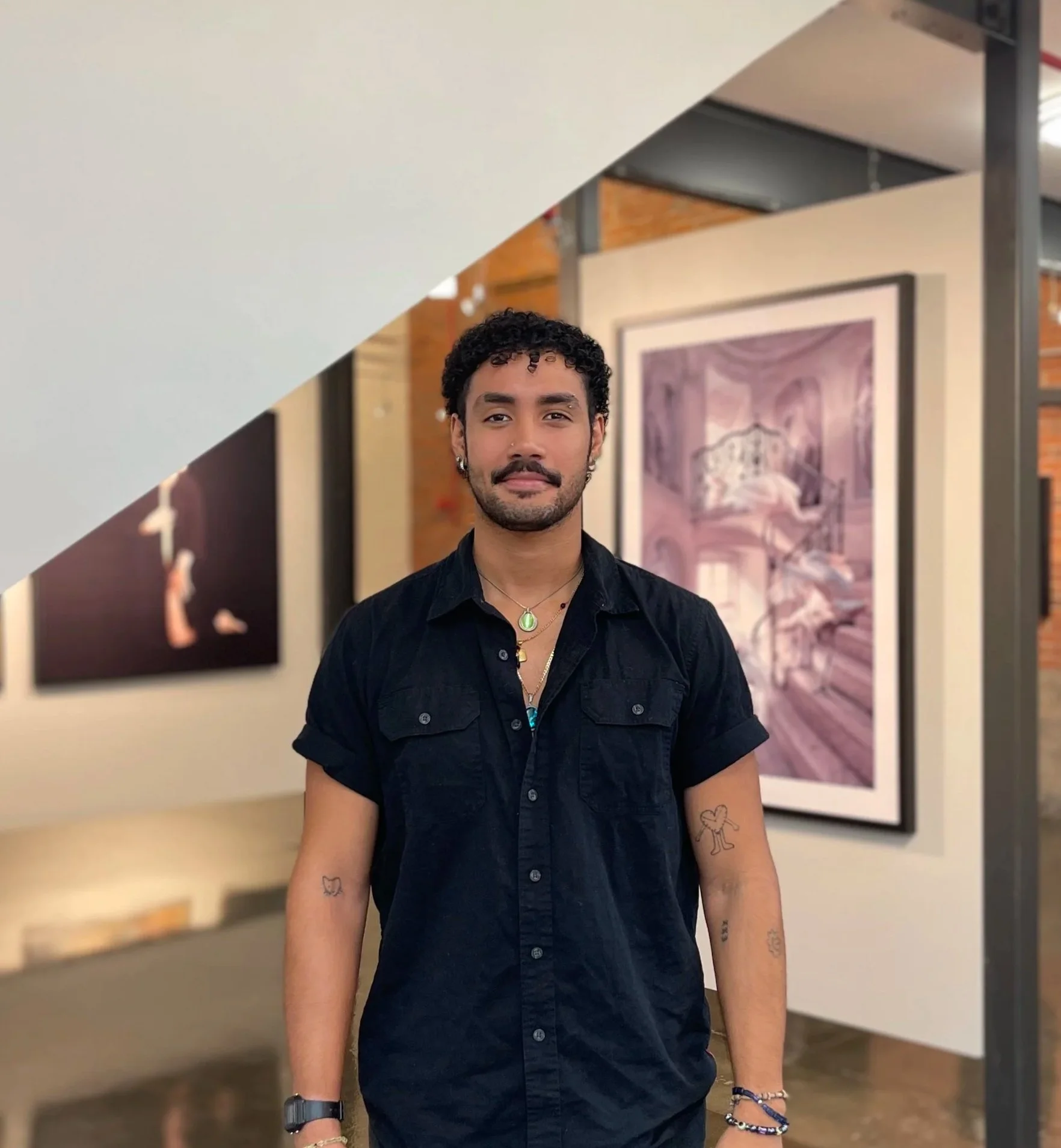 A man with short curly hair, a mustache, and earrings standing indoors in front of framed artwork. He is wearing a black button-up shirt, a necklace, a watch, and multiple bracelets, with tattoos on his arms.