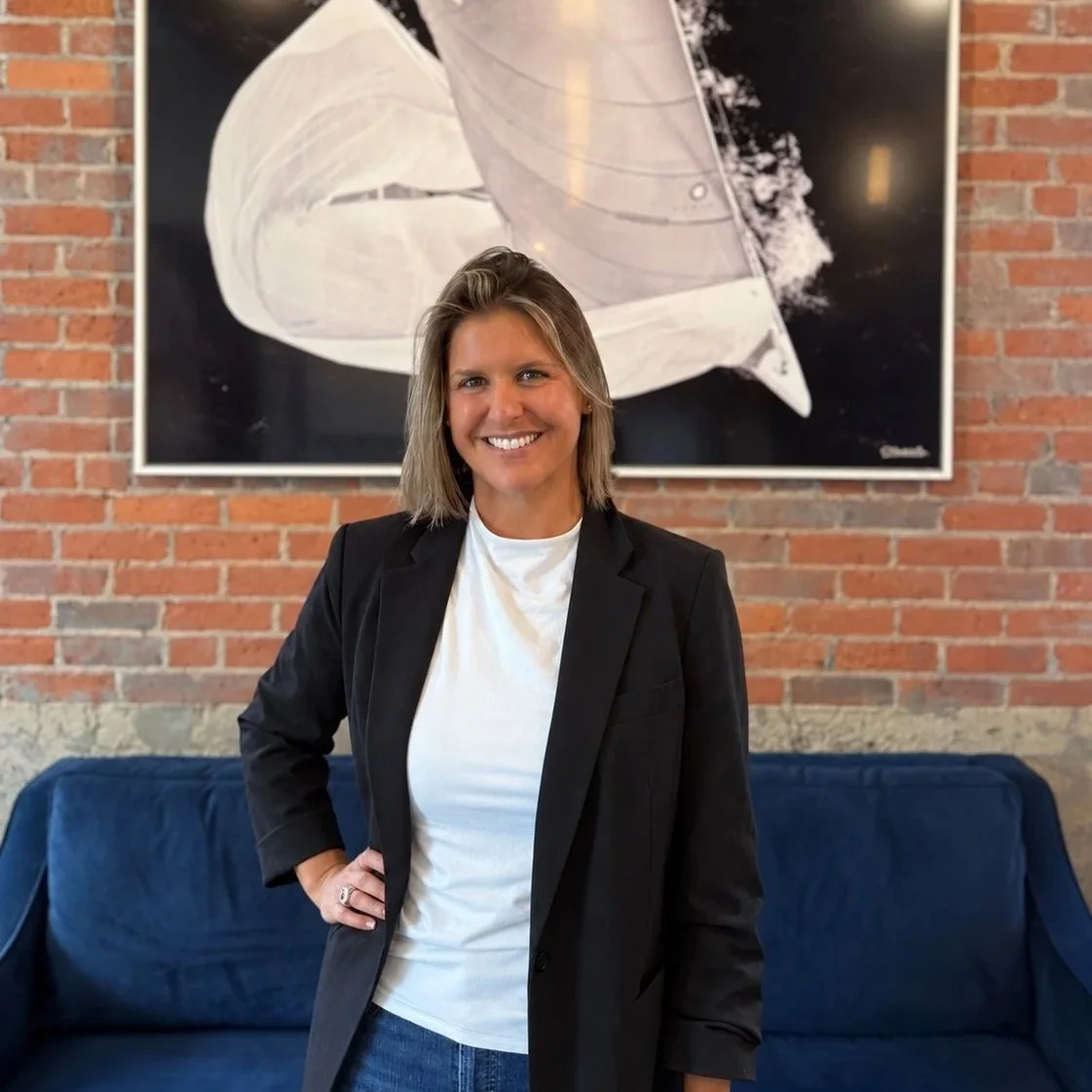 Smiling woman in black blazer and white t-shirt standing in front of a brick wall with modern art painting behind her