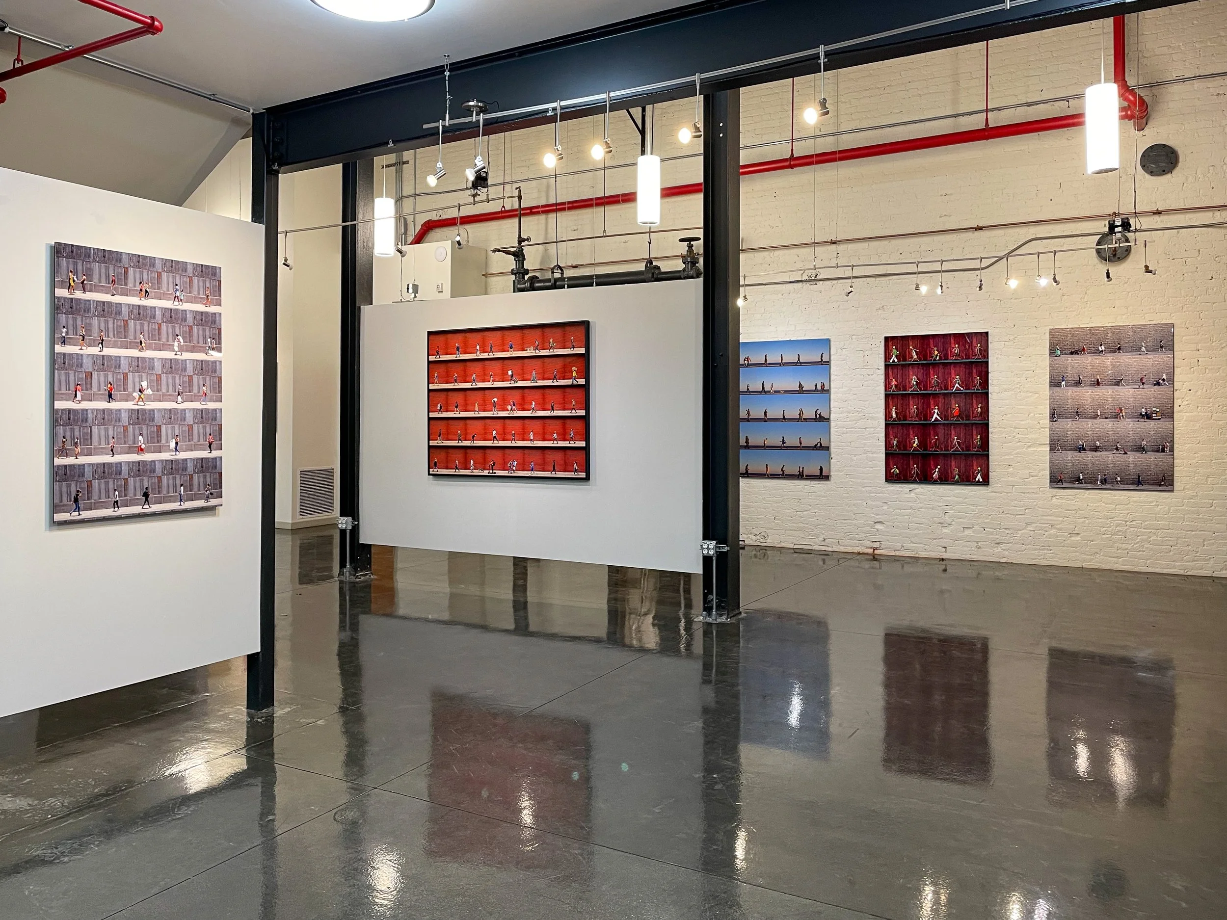 Art gallery with displayed art pieces of figures walking along different backgrounds, including a red stage, blue sky, and brick wall, with gallery lights illuminating the artwork.