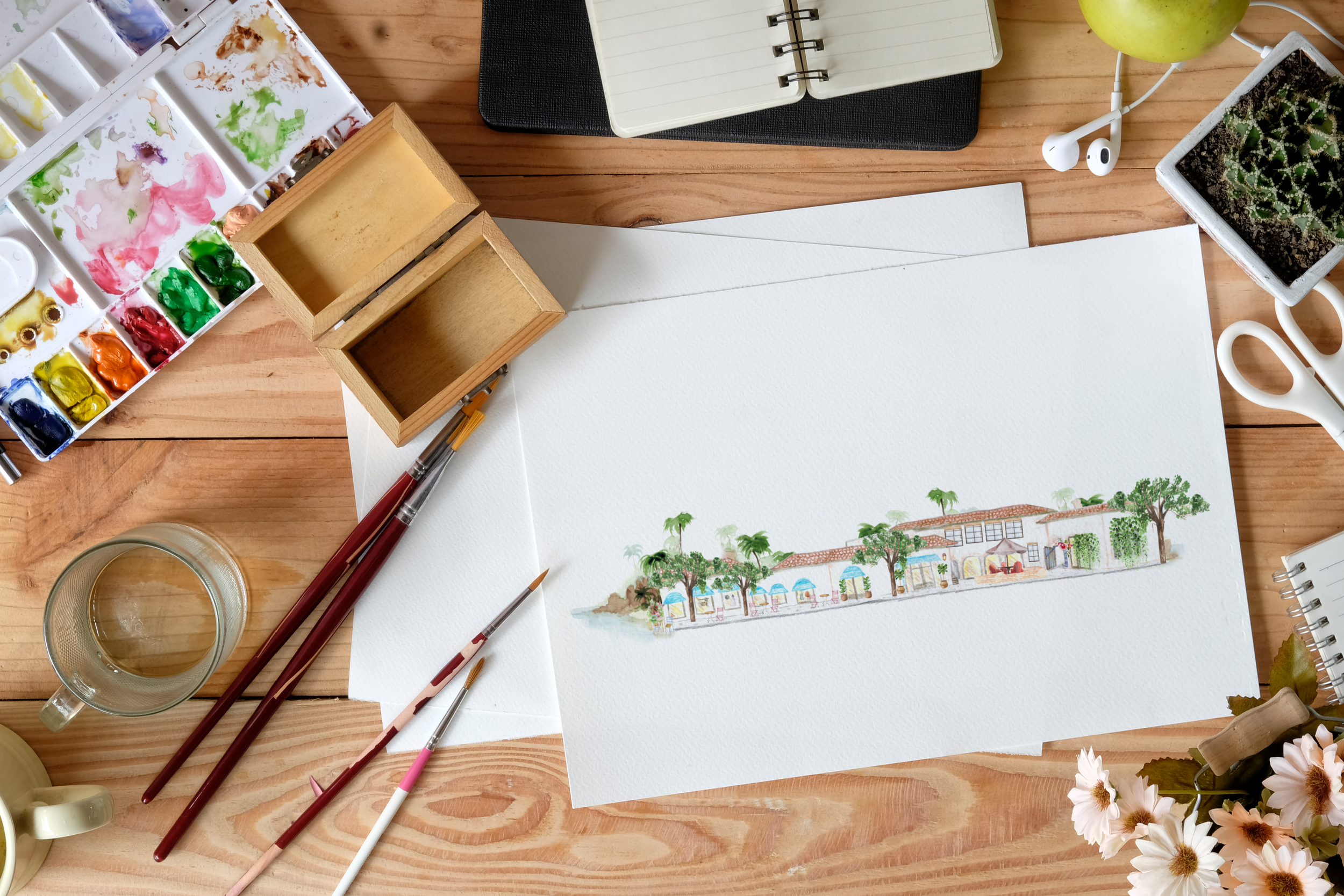 Behind-the-scenes artist workspace showing the hand-illustrated Plaza Del Sol building created for the Wendy Foster destination map, surrounded by watercolor supplies, brushes, and creative tools used throughout the illustration process.