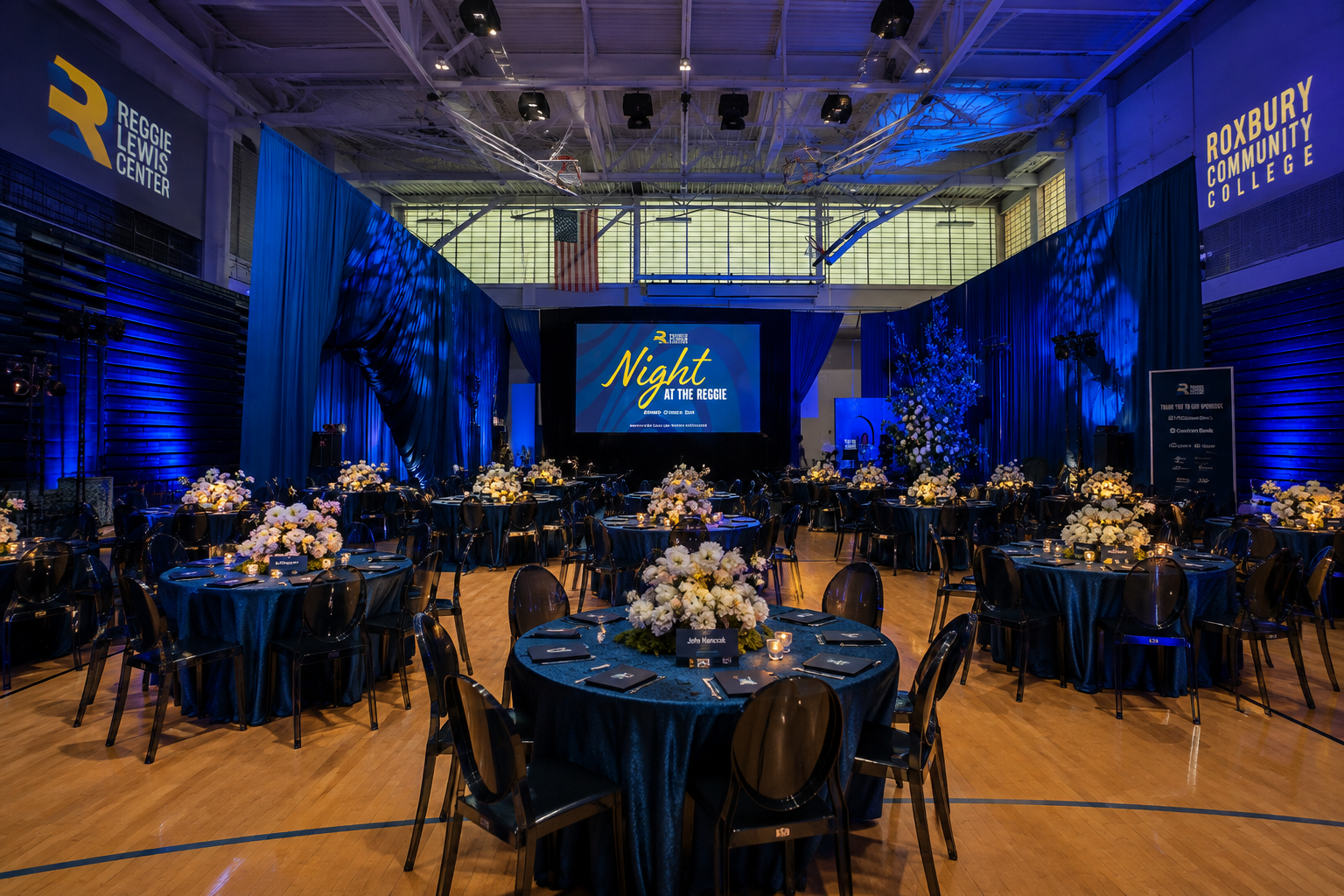 Night at the Reggie event inside a transformed gymnasium, with deep blue draping, dramatic lighting, branded stage graphics and elegant tables showing how event design can work with an existing venue’s architecture.