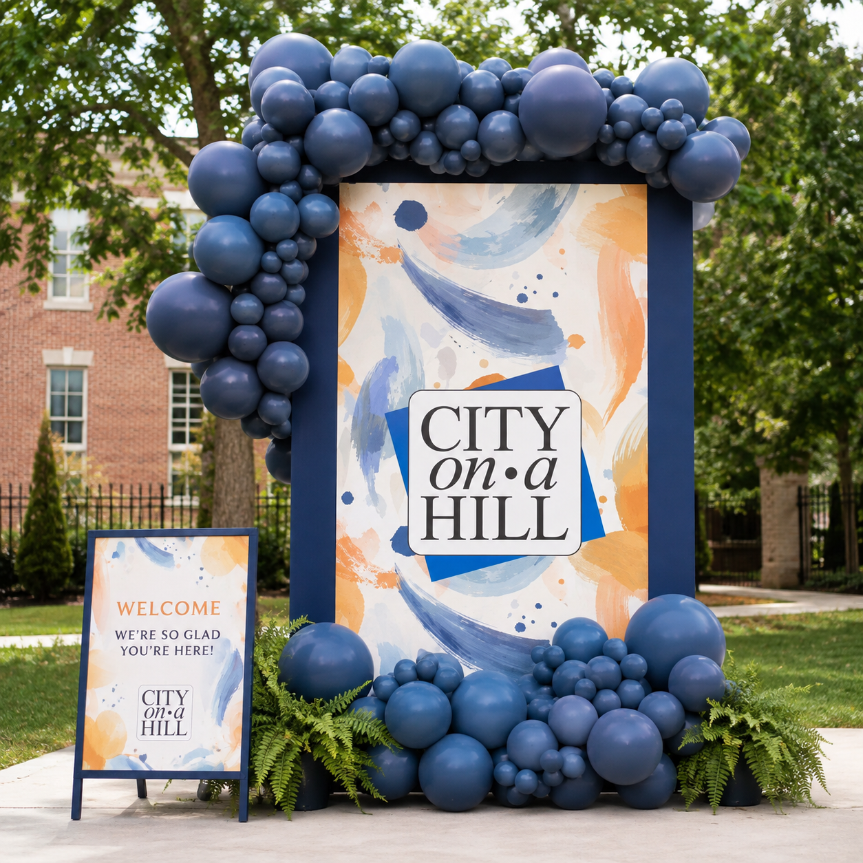 Large outdoor welcome sign featuring a custom abstract pattern in the City on a Hill brand colors, framed by an organic navy balloon installation for a community event entrance.