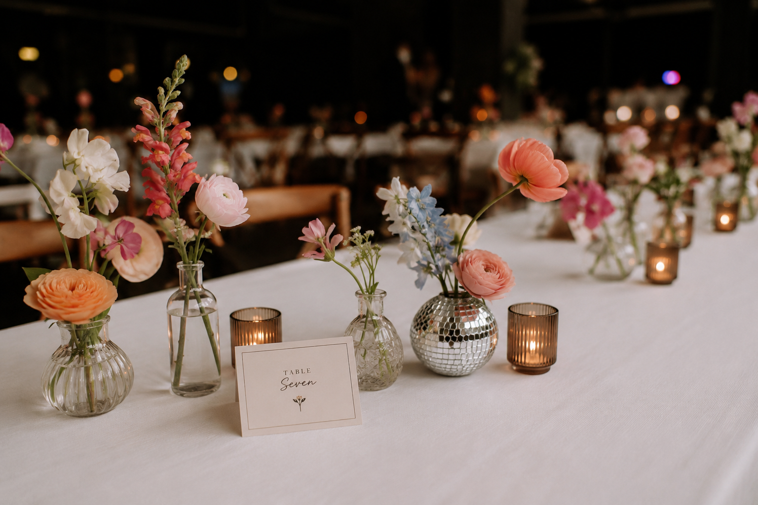 Airy wedding reception table with colorful bud vase florals, amber candlelight, a mirrored disco ball vase and simple table stationery.