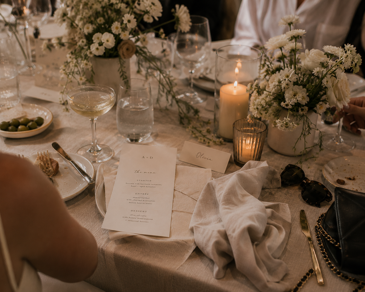 Lived-in wedding reception table with menus, place cards, half-finished cocktails, candles, white florals, rumpled linen napkins, sunglasses and a handbag, showing how thoughtful event branding becomes part of the guest experience.