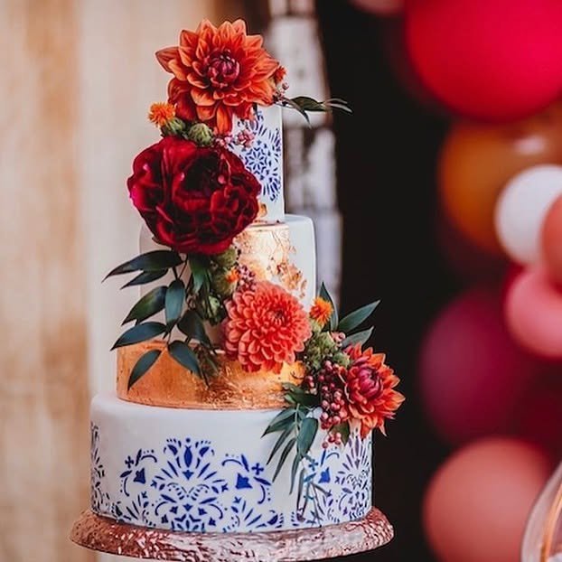 Reference photo of an elegant tiered wedding cake with hand-painted blue tile details, gold accents, and vibrant red and orange florals, created to be illustrated and incorporated into the couple's custom wedding menu design.