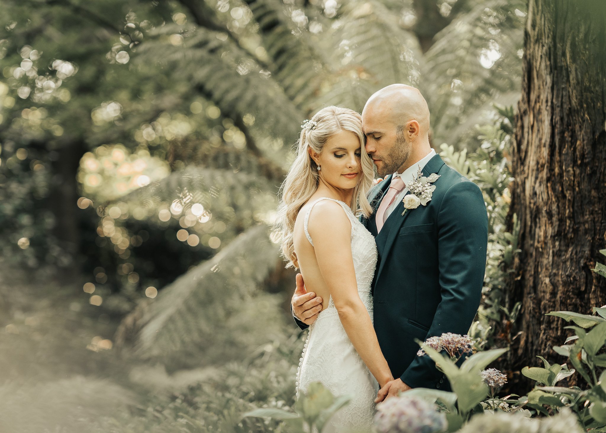 A bride and groom share an intimate moment in a lush, green outdoor setting surrounded by trees and foliage. The bride has long, blonde hair and wears a white wedding gown, while the groom is bald, dressed in a dark suit with a boutonnière, and gently holds the bride's waist.