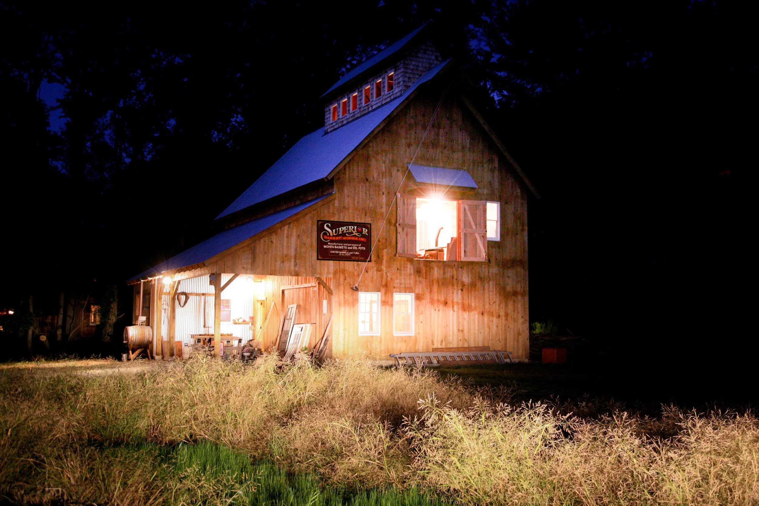 Wooden barn illuminated at night with an open door and a visible ladder, surrounded by tall grass.