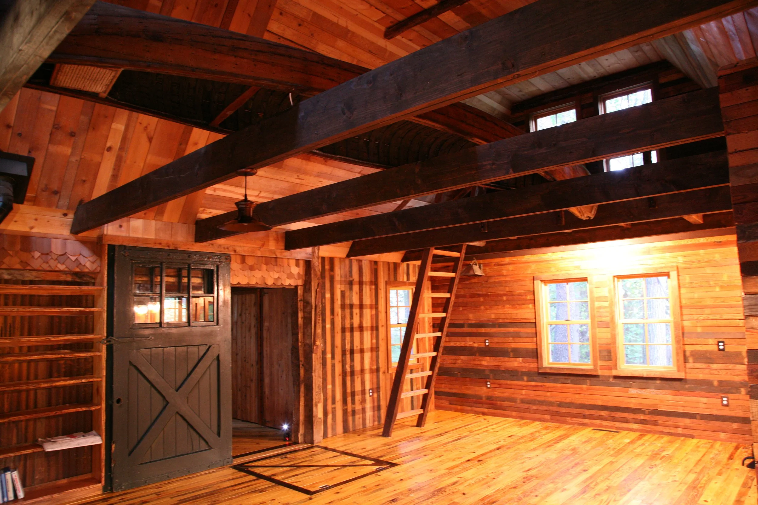 Interior of a rustic wooden cabin with exposed beams, wood-paneled walls, ladder to loft area, and a large window.