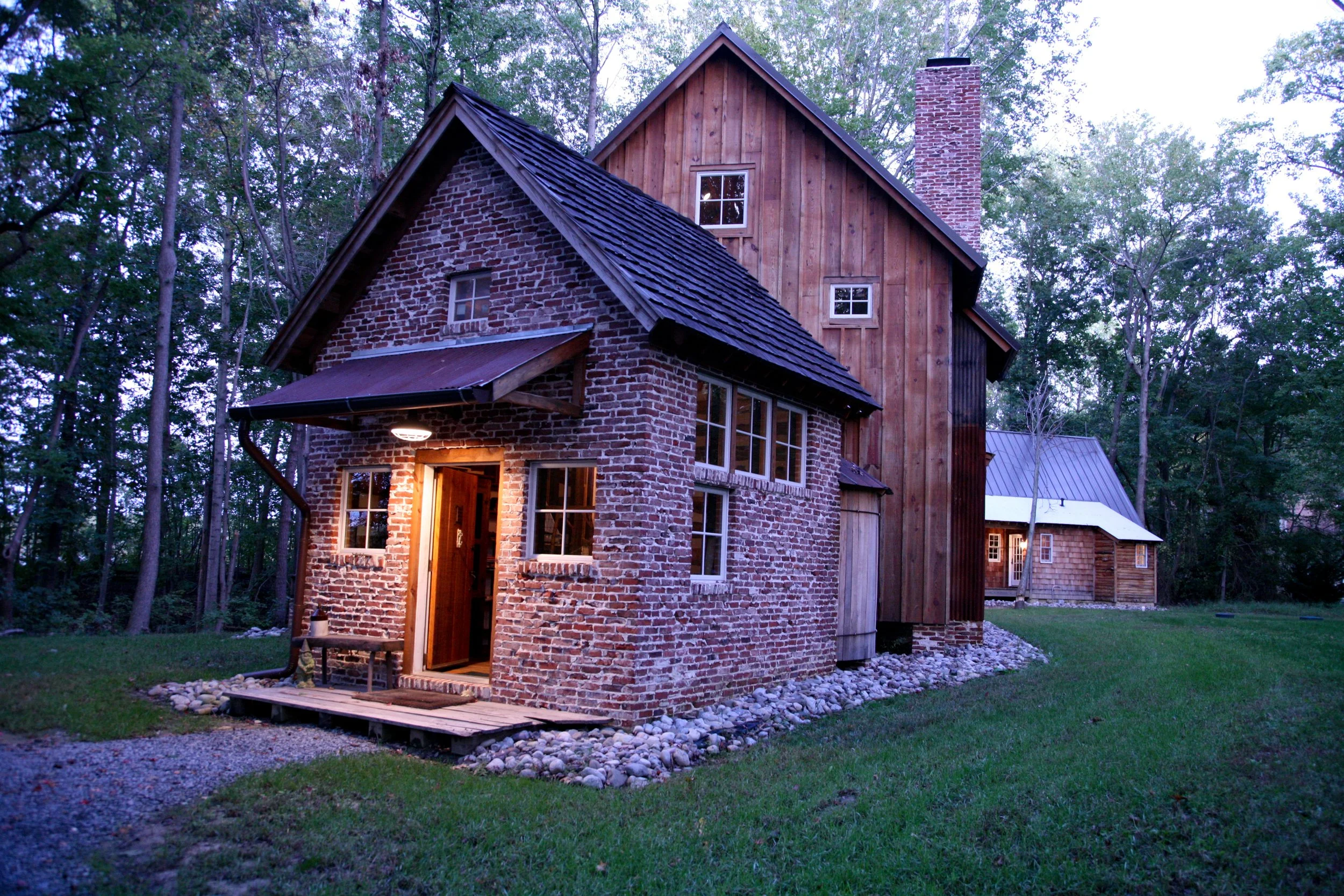 Brick and wood house in a wooded area, lit by exterior lights, with a smaller wooden structure nearby, surrounded by trees.
